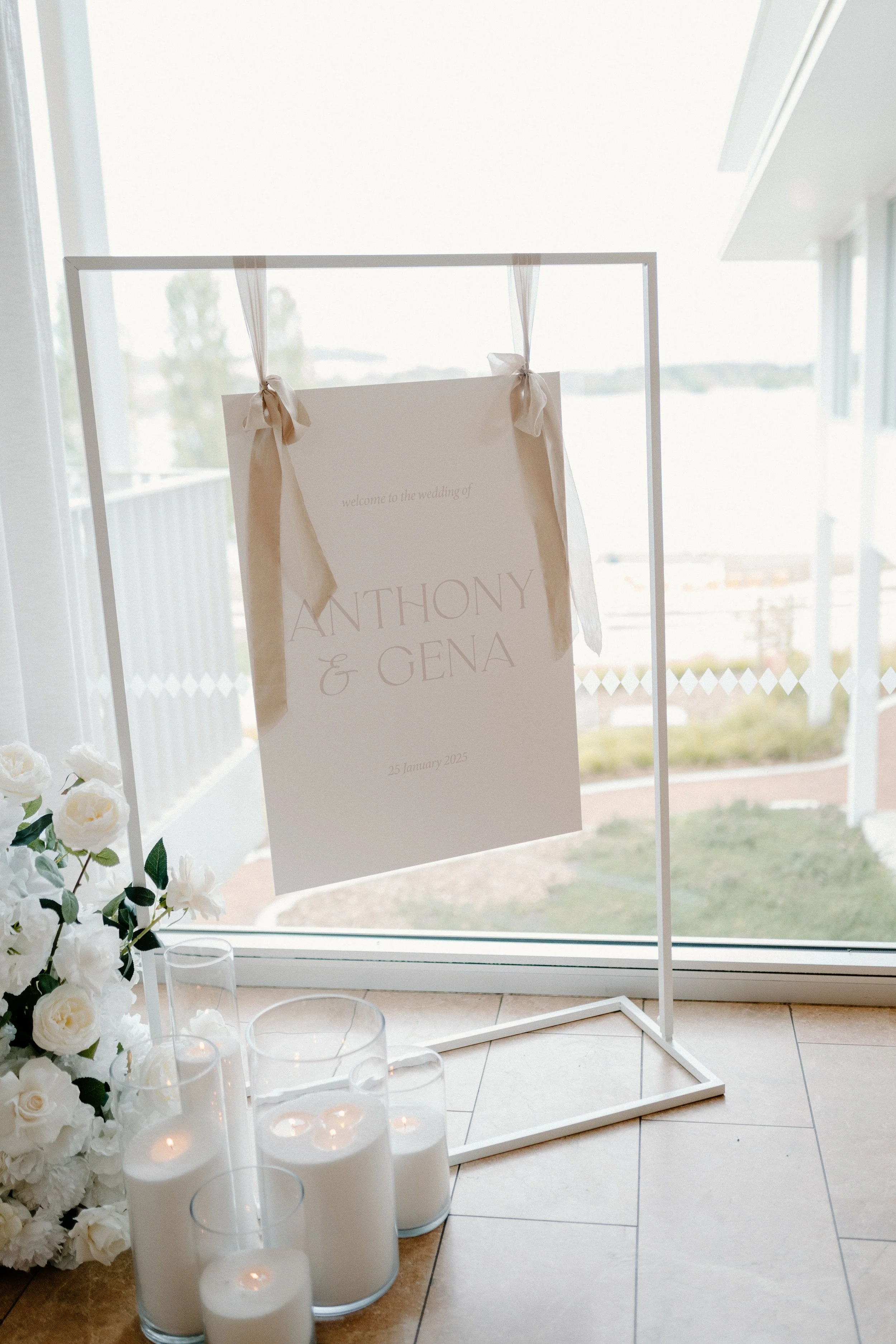 Wedding welcome sign with beige ribbons hanging on a white frame, surrounded by white flowers and candles, with a large window and balcony in the background.