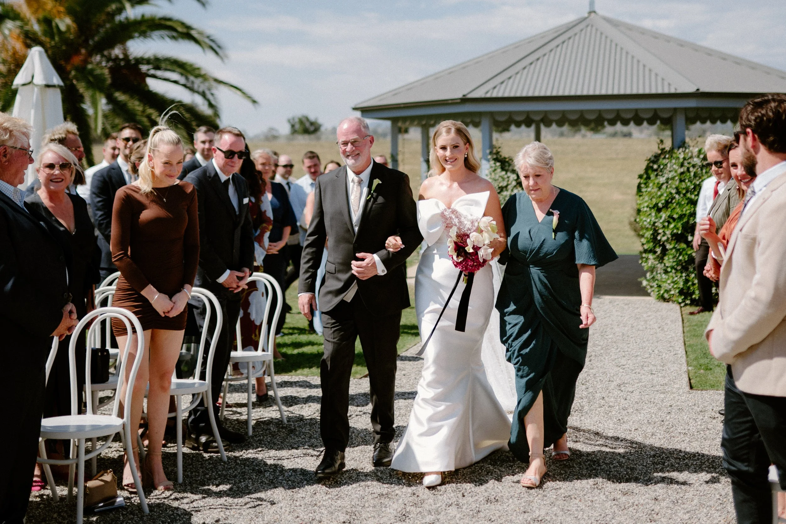 Bride walking down the aisle with her father at an outdoor wedding ceremony, surrounded by guests.