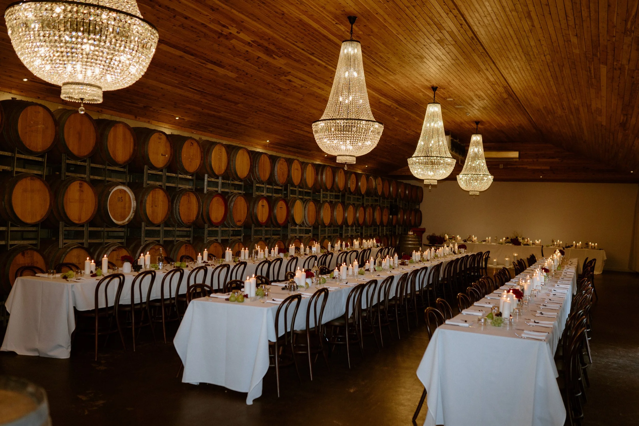 The Sienna, Hunter Valley Wedding Venue, Wedding Photos. A banquet hall decorated for a special event with long tables covered in white tablecloths, candles, and floral centerpieces, against a wall of wine barrels and illuminated by large crystal cha
