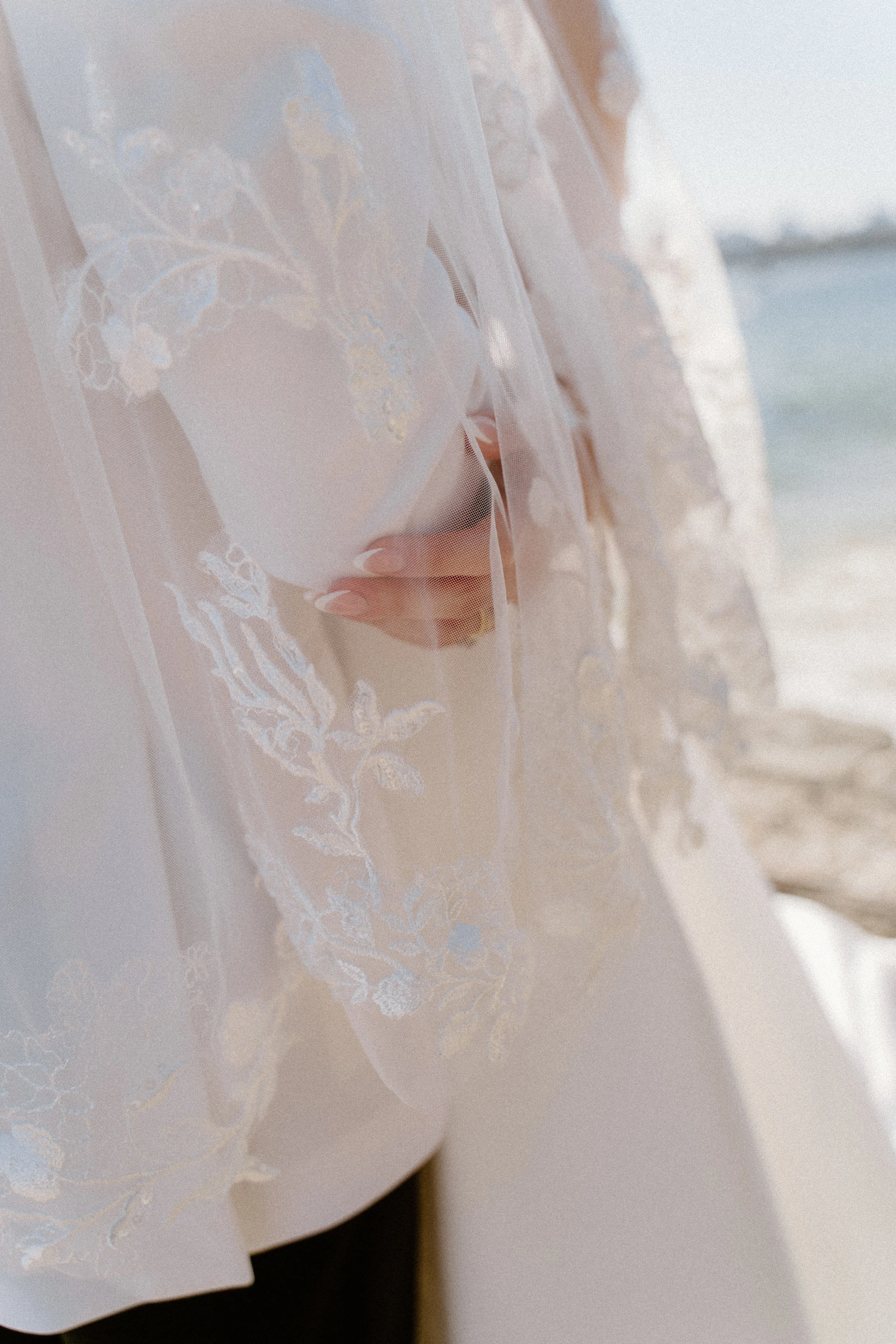 A bride holding a bouquet, partially hidden by a delicate lace veil, near a beach with water and sky in the background.