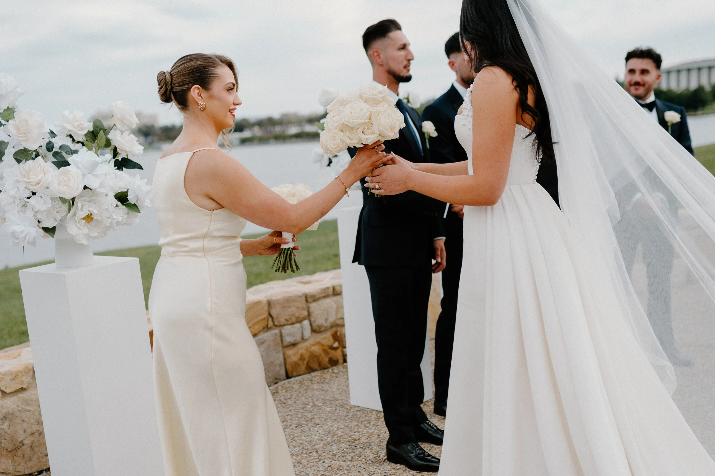 A wedding ceremony outdoors by a river with a city in the background, featuring a bride in a white wedding gown and veil, a woman in a cream dress handing her a bouquet, and three men in black suits and bow ties.