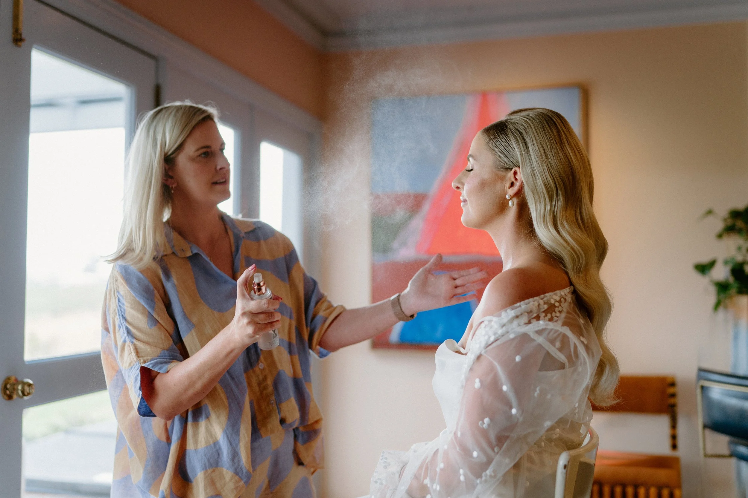 A woman is applying makeup to another woman, who is sitting and smiling during her beauty routine in a room with natural light from large windows.