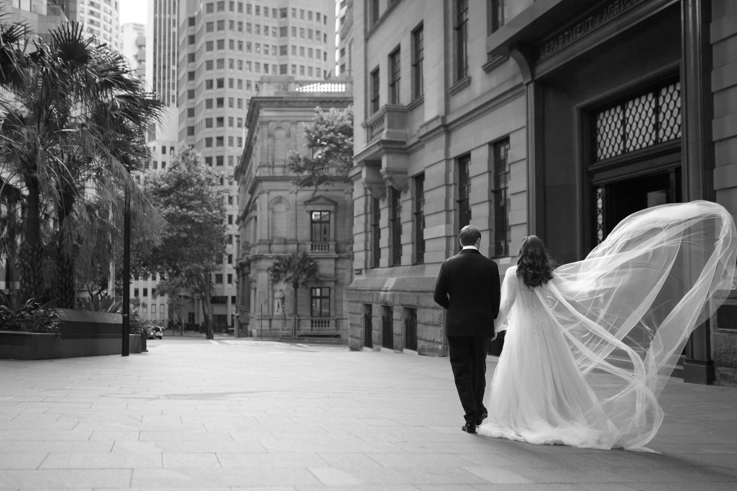A bride and groom walking together on a city street, with the bride's long veil flowing in the wind.