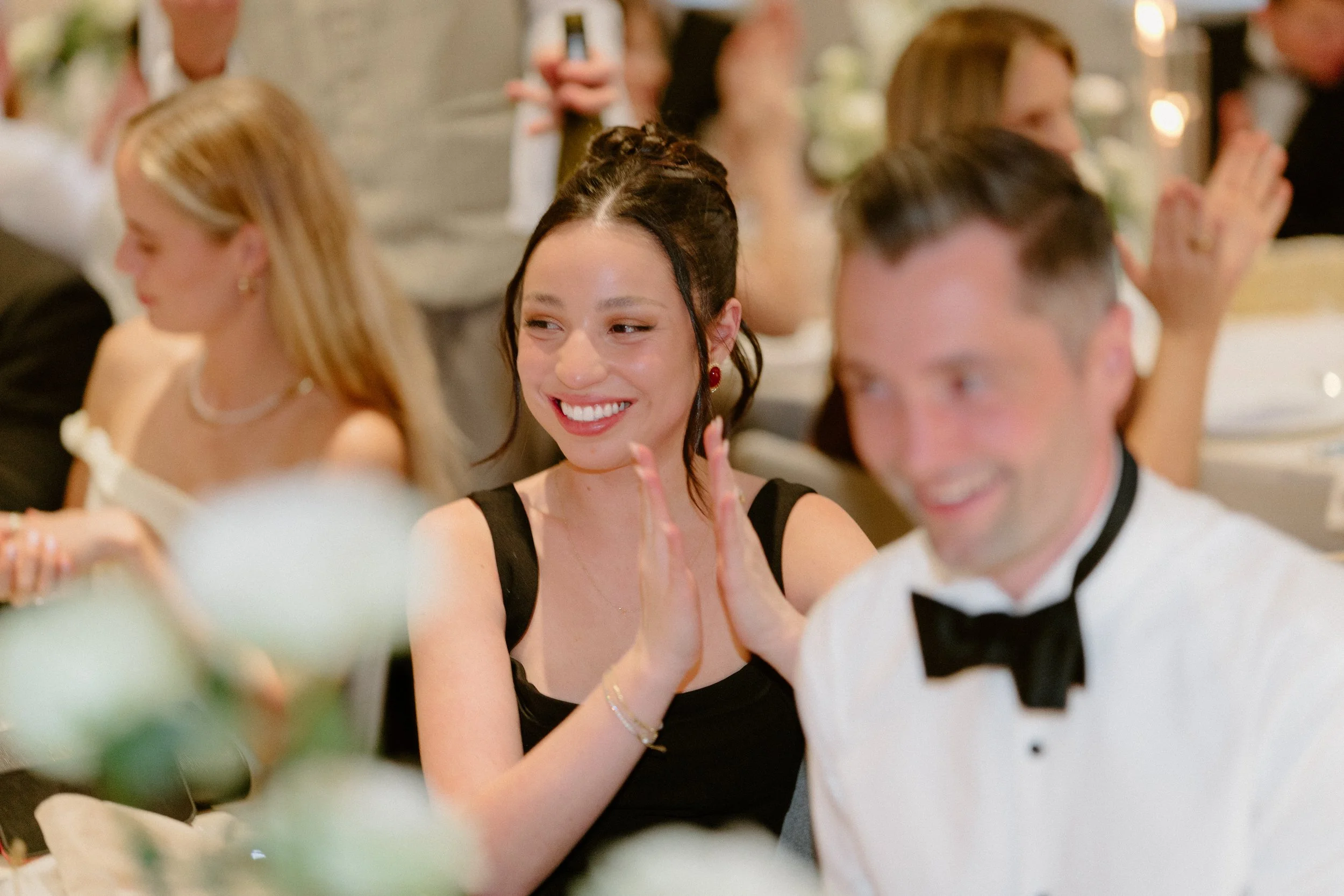 People sitting at a formal event, with one woman smiling and clapping, wearing a black dress and red earrings, and a man in a tuxedo with a bow tie blurred in the foreground.