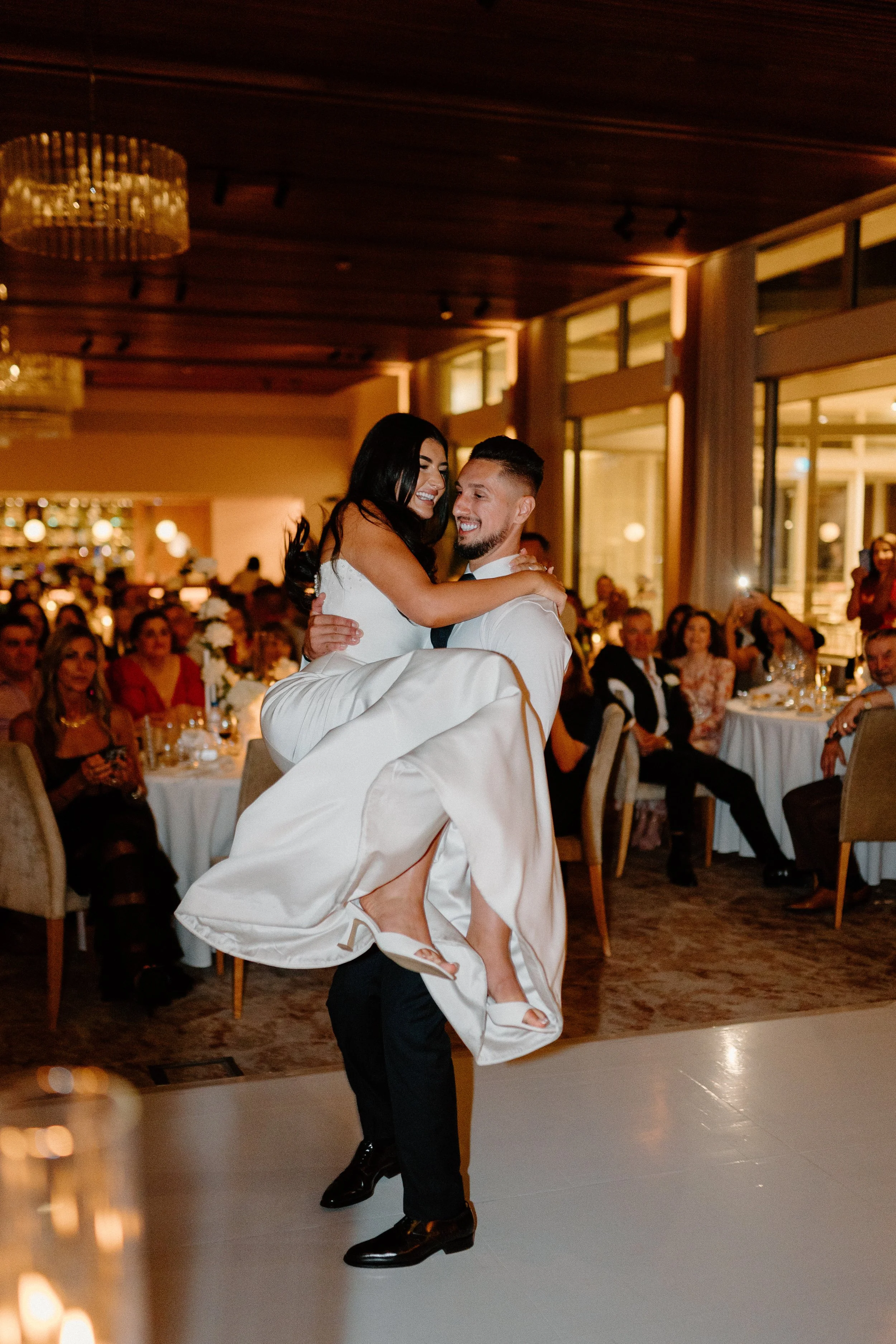 Newlywed couple dancing at wedding reception, the groom lifting the bride on the dance floor with guests seated at tables in the background.