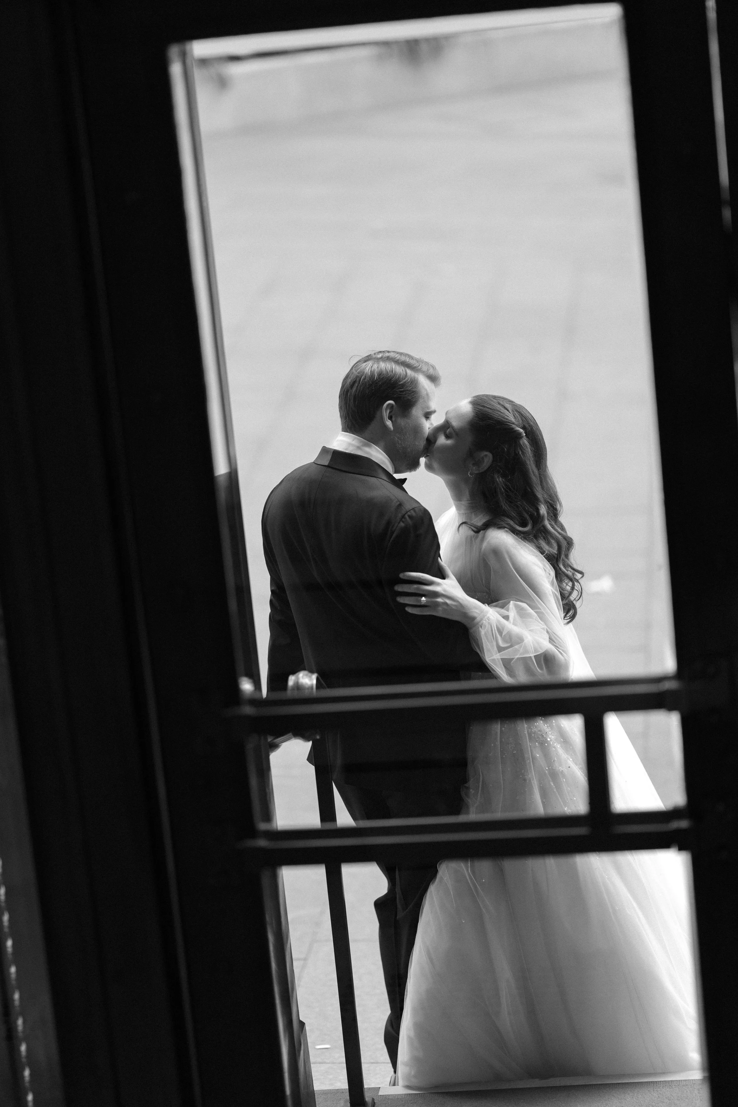A black and white photo of a bride and groom sharing a kiss, seen through a window.
