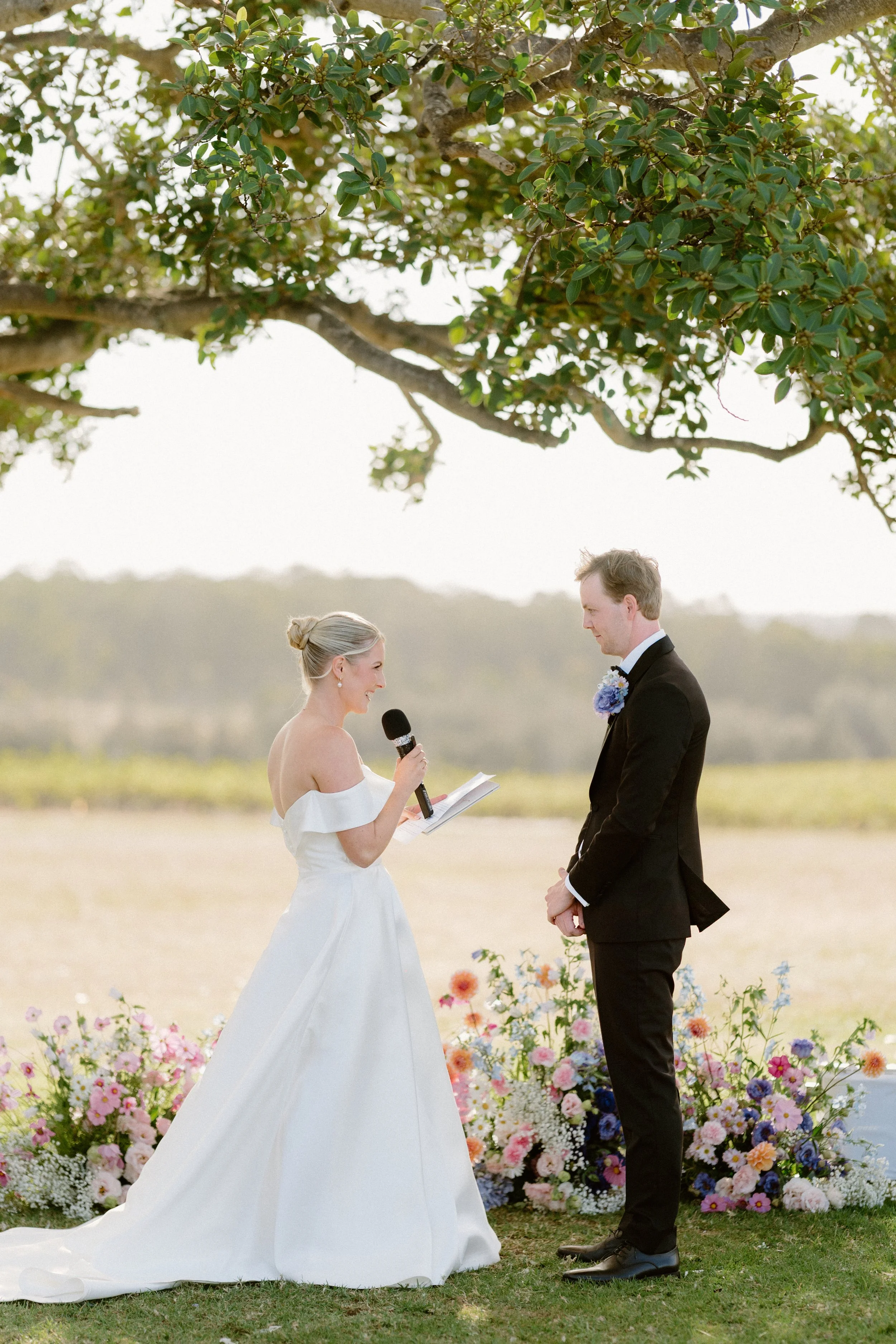 A bride and groom exchanging vows during an outdoor wedding ceremony under a tree, with colorful flowers arranged at their feet. Bimbadgen Palmers Lane Wedding, Hunter Valley
