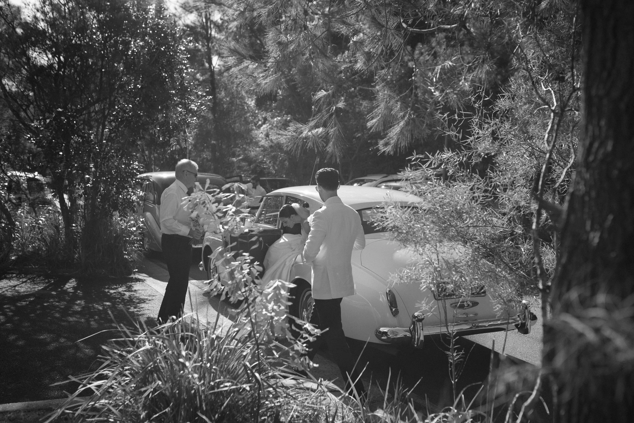 Black and white photo of a bride and groom getting into a vintage car, with an older man holding flowers nearby, surrounded by trees and parked cars, during daytime.