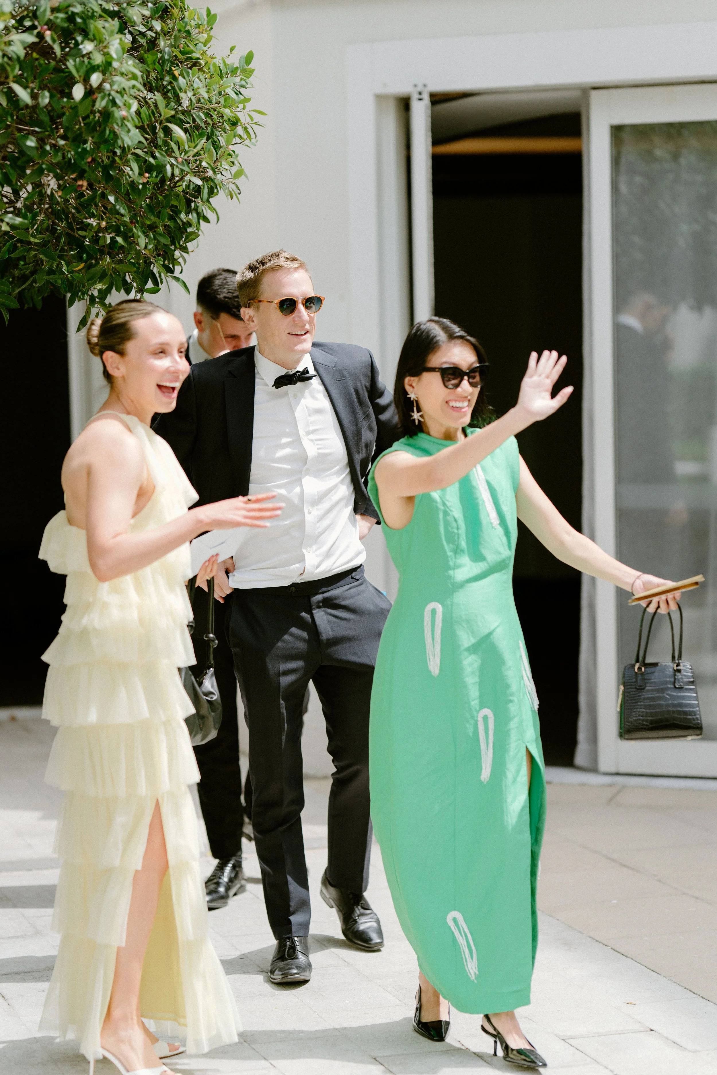 Group of four people smiling and waving outside a building, dressed in formal and semi-formal attire.