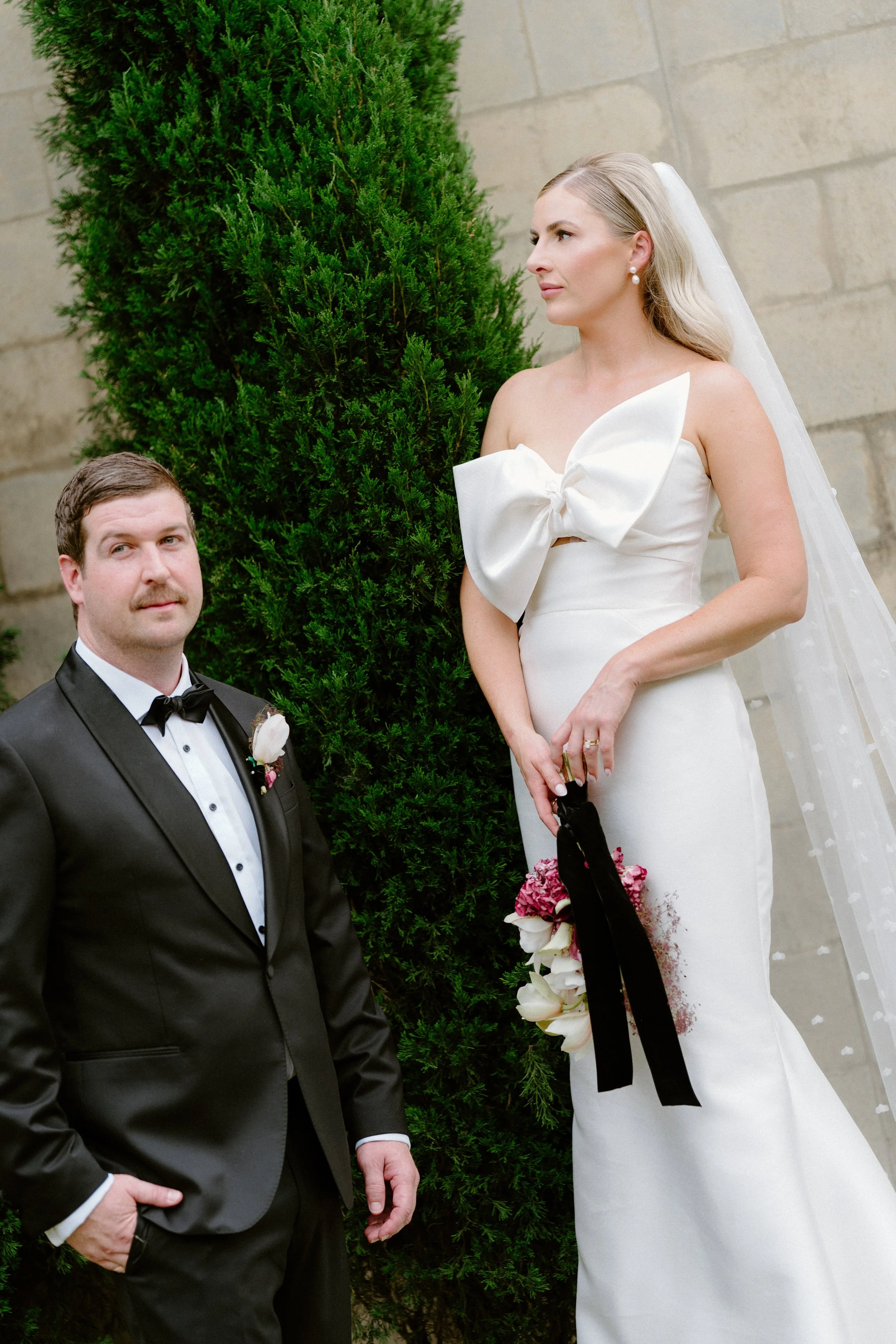 Bride in a white wedding dress with a large bow on the bodice, holding a bouquet of white and pink flowers, standing next to a groom in a black tuxedo with a white shirt and black bow tie, outdoors near a tall green shrub and stone wall.