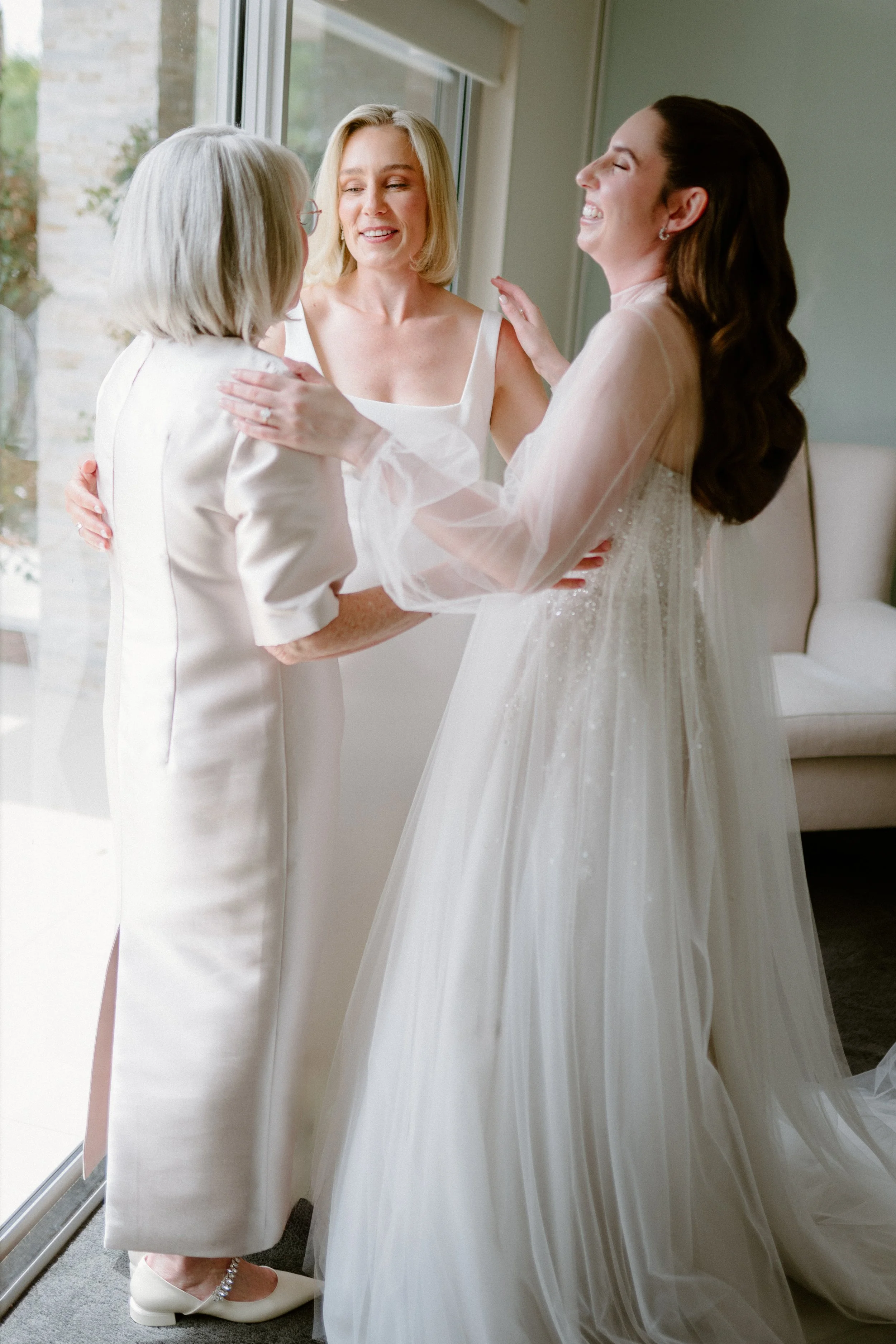 Three women, one in a wedding gown, are happily embracing in a bright room near a large window.