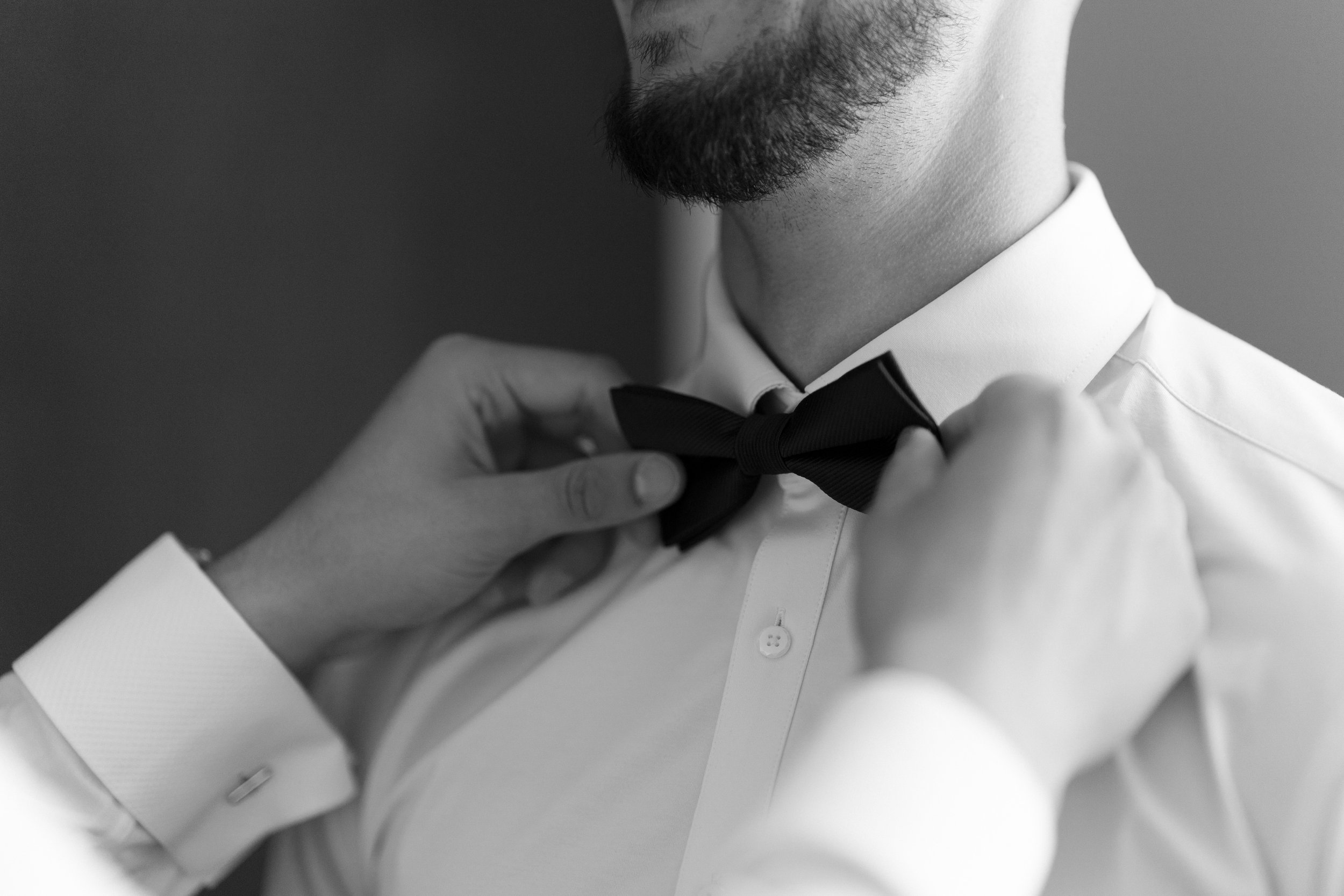 A man adjusting a black bow tie, wearing a white dress shirt with French cuffs. The image is in black and white.