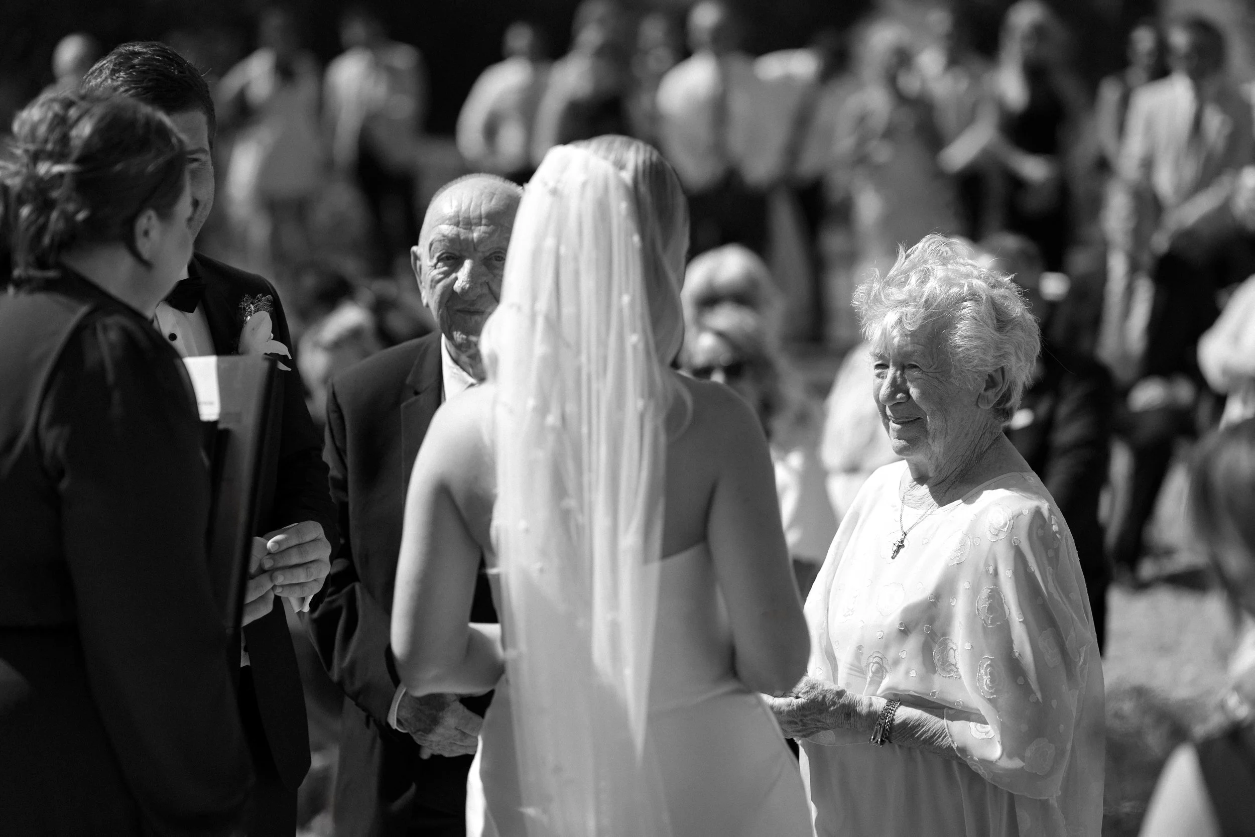 A bride and an elderly woman talking at a wedding, with other guests in the background.