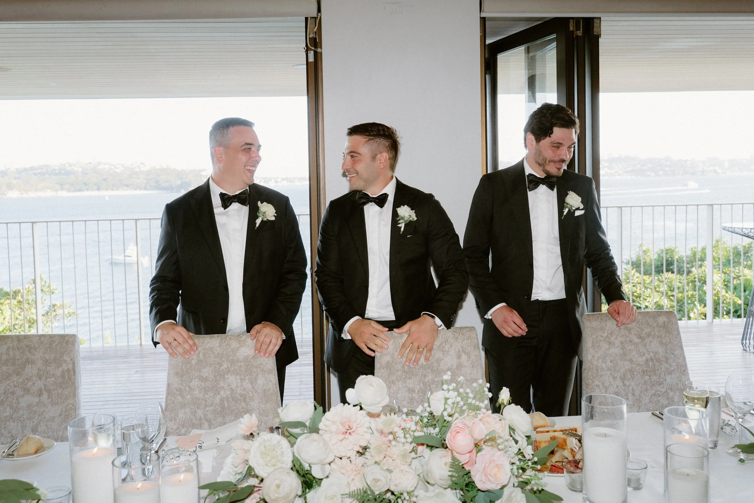Three men in tuxedos with white shirts and black bow ties standing around a banquet table with a floral centerpiece, in front of a large window overlooking a body of water, during a wedding celebration.