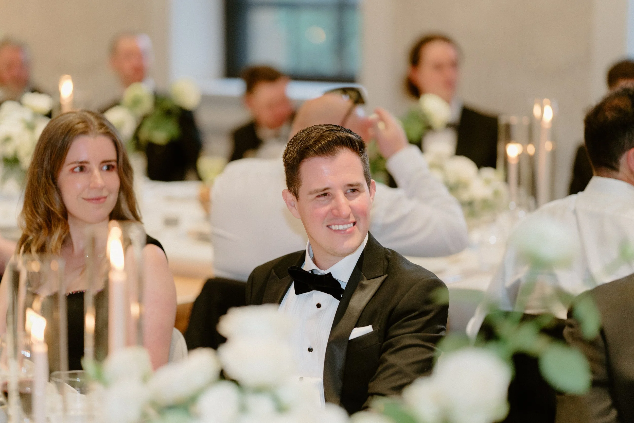 A man in a tuxedo smiling and sitting at a formal event, surrounded by guests in elegant attire, with floral decorations and candles on the table.