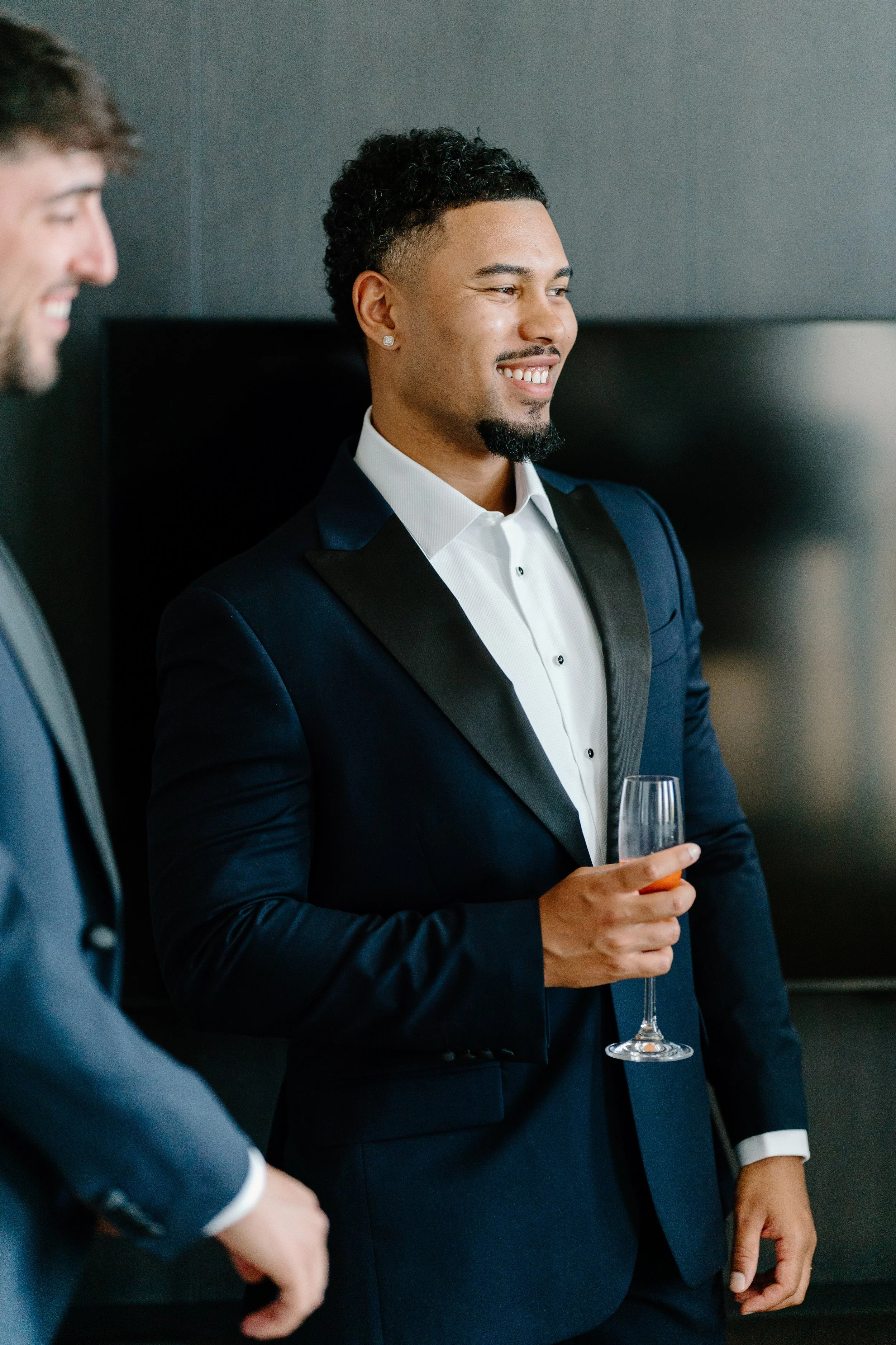 A man dressed in a tuxedo holding a wine glass at a formal event, smiling and engaged in conversation.