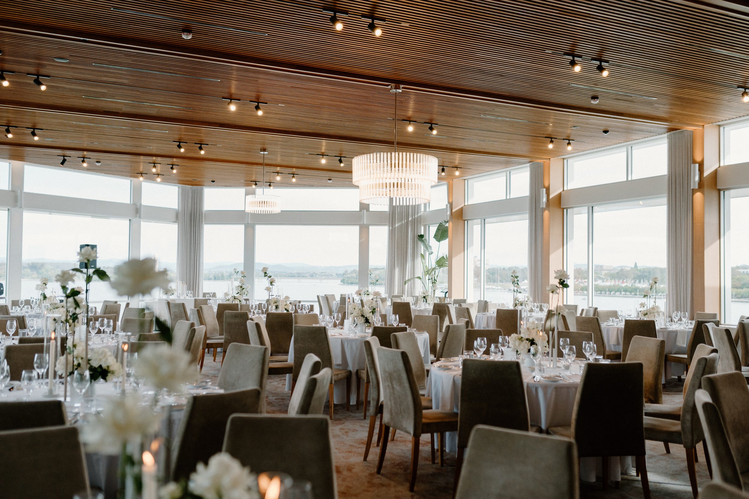 Elegant banquet hall with large windows, round tables covered with white tablecloths, decorated with white floral centerpieces, set with glassware and candles, and modern chandeliers hanging from the wooden ceiling.