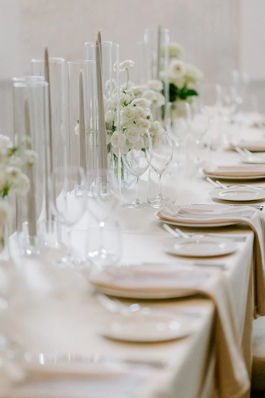 Elegant dining table setup with white tablecloth, white floral centerpieces, clear glassware, and plates with napkins.