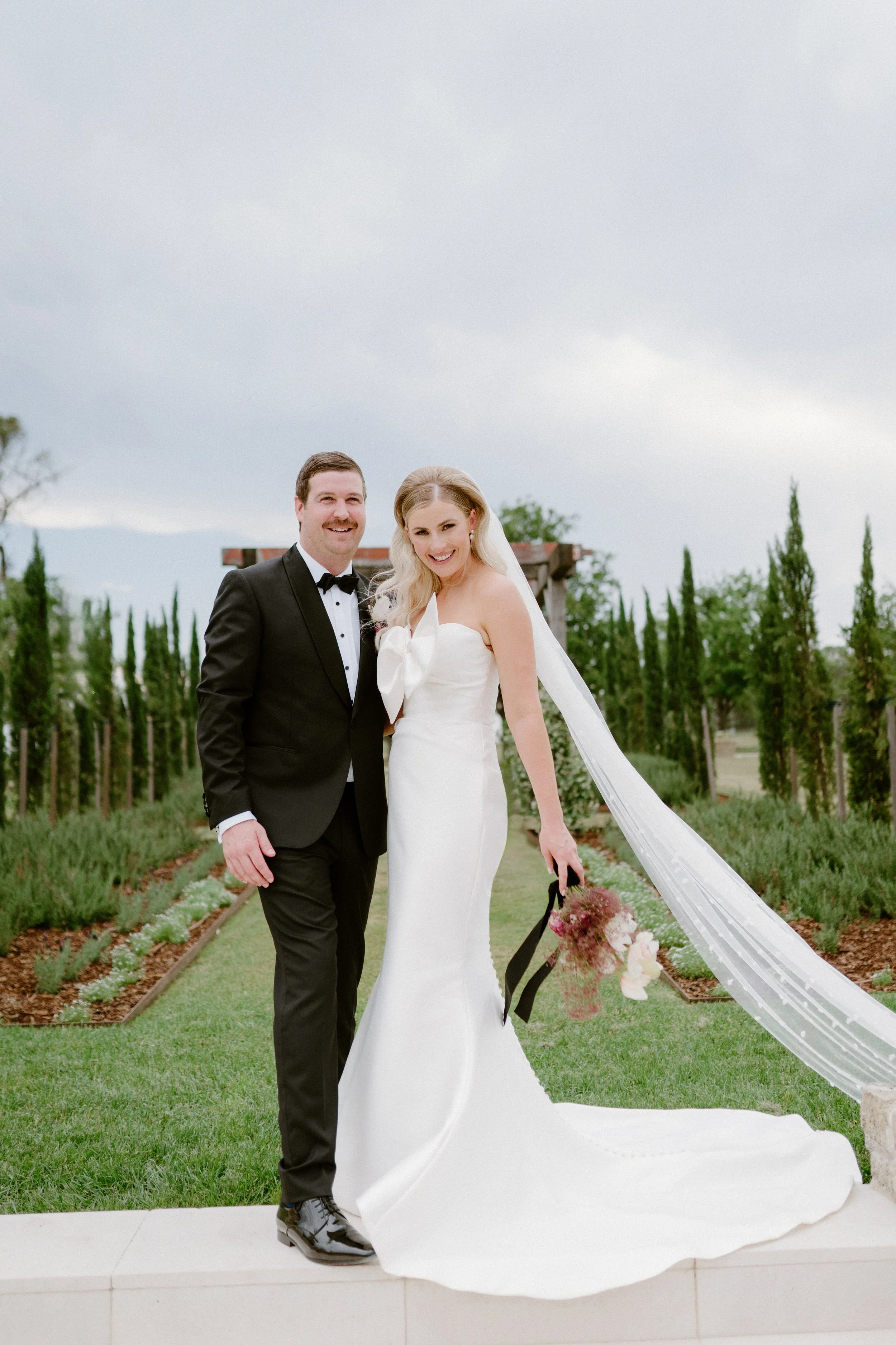 A newlywed couple stands outdoors on a cloudy day, dressed in wedding attire. The groom is in a black tuxedo with a bow tie, and the bride is in a strapless white wedding gown holding a bouquet of pink and white flowers. They are smiling and posing f