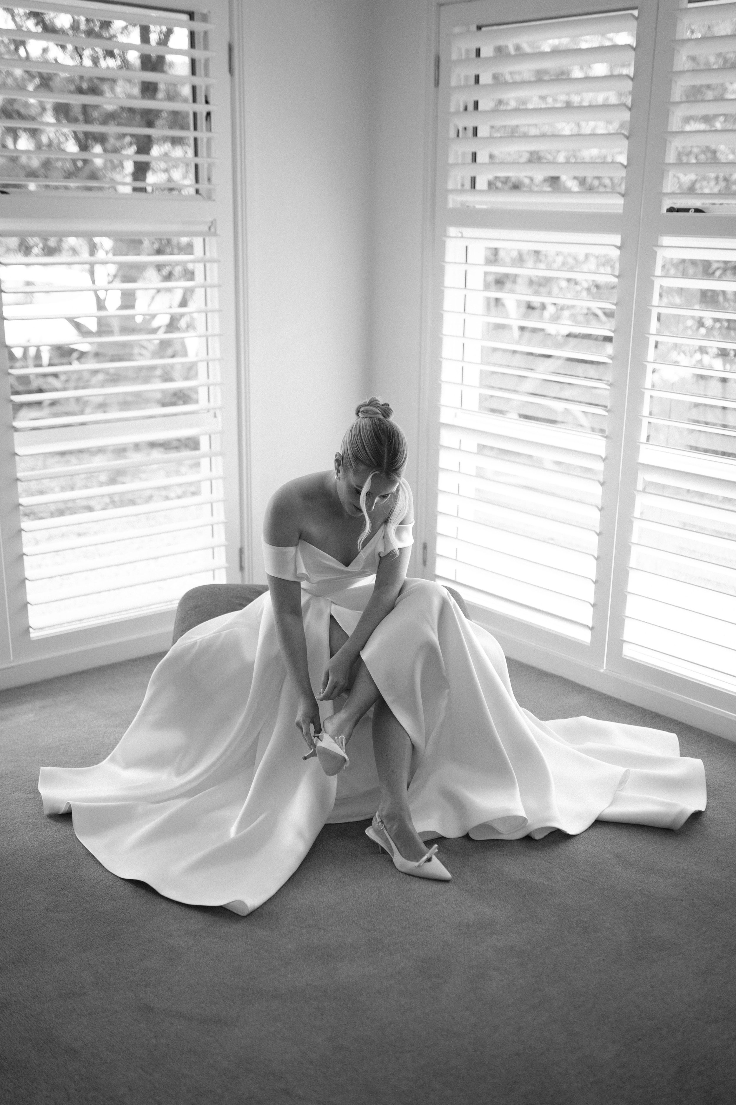A bride in a strapless wedding dress sitting on a cushioned stool, putting on her high-heeled shoe in a room with large windows and shutters.