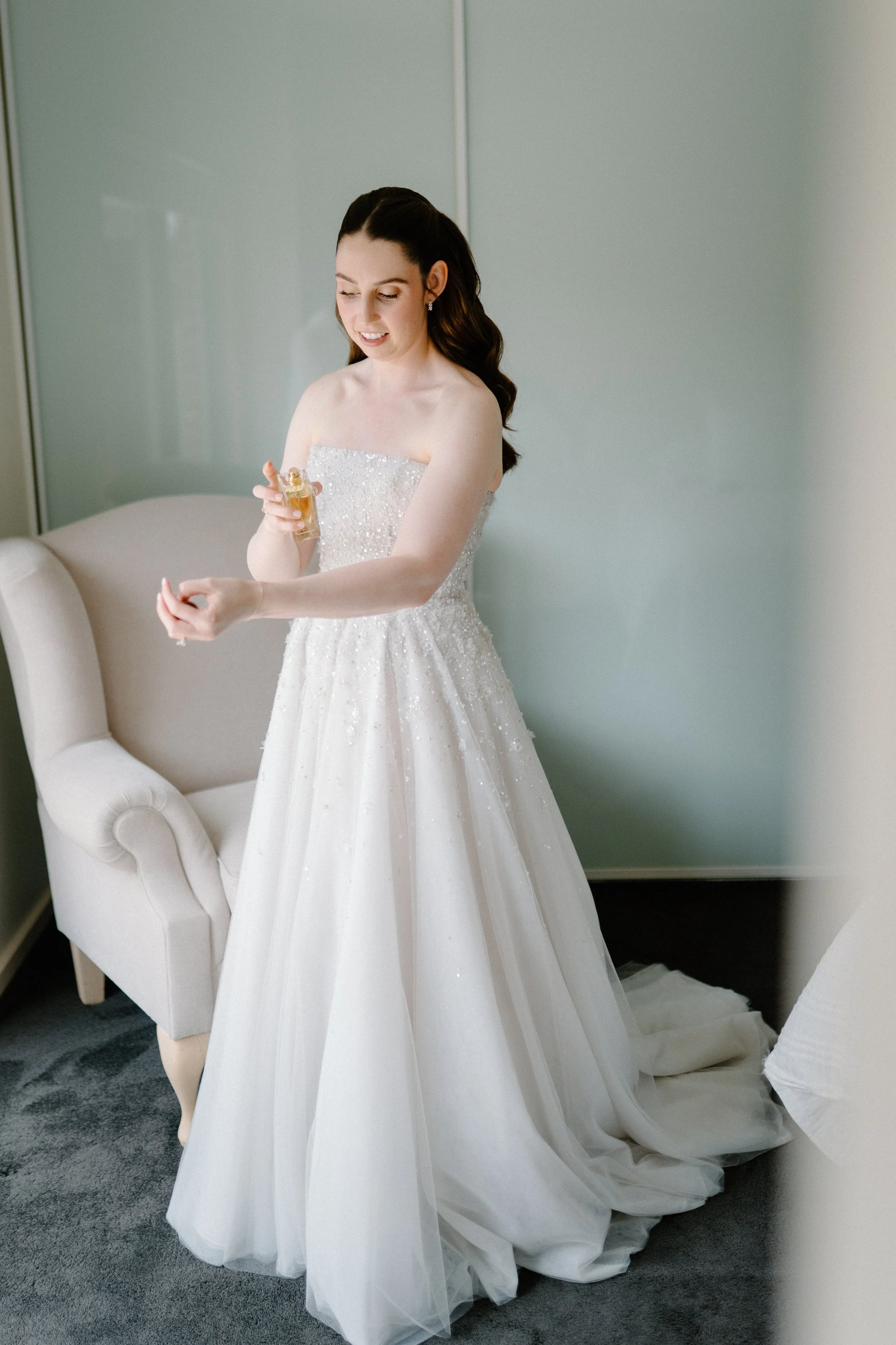 Bride in a strapless white wedding gown applying perfume in a softly lit room.