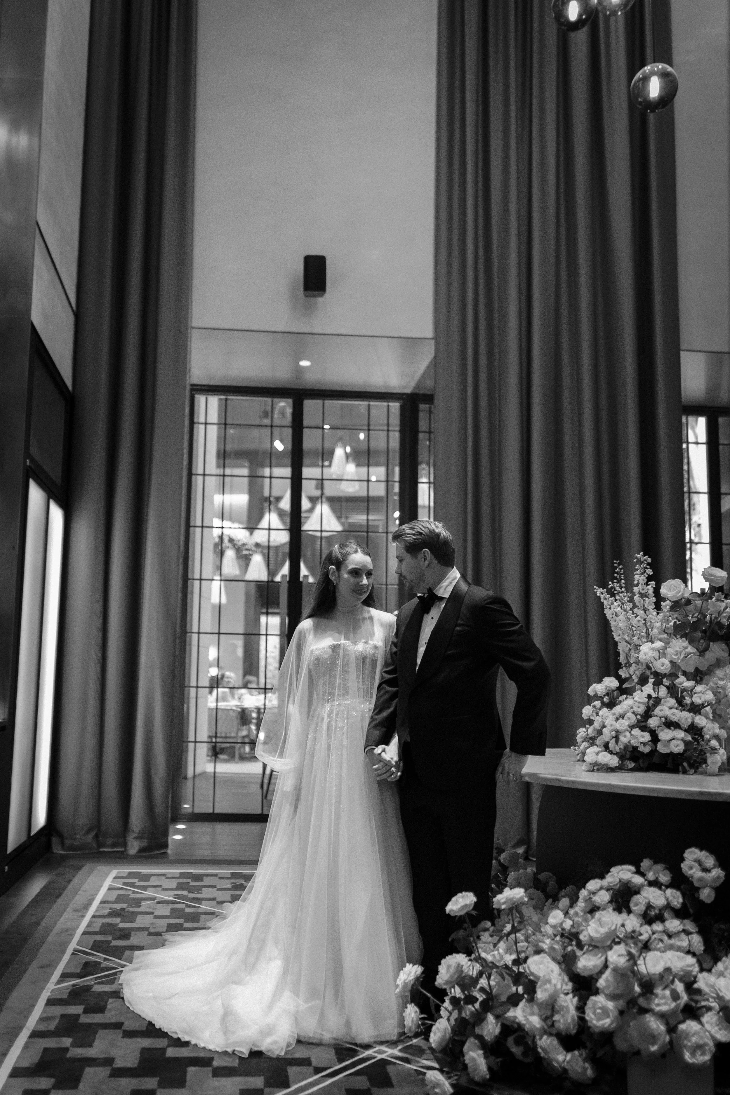 A black and white photo of a bride and groom holding hands and looking at each other inside a decorated venue with large windows and curtains.