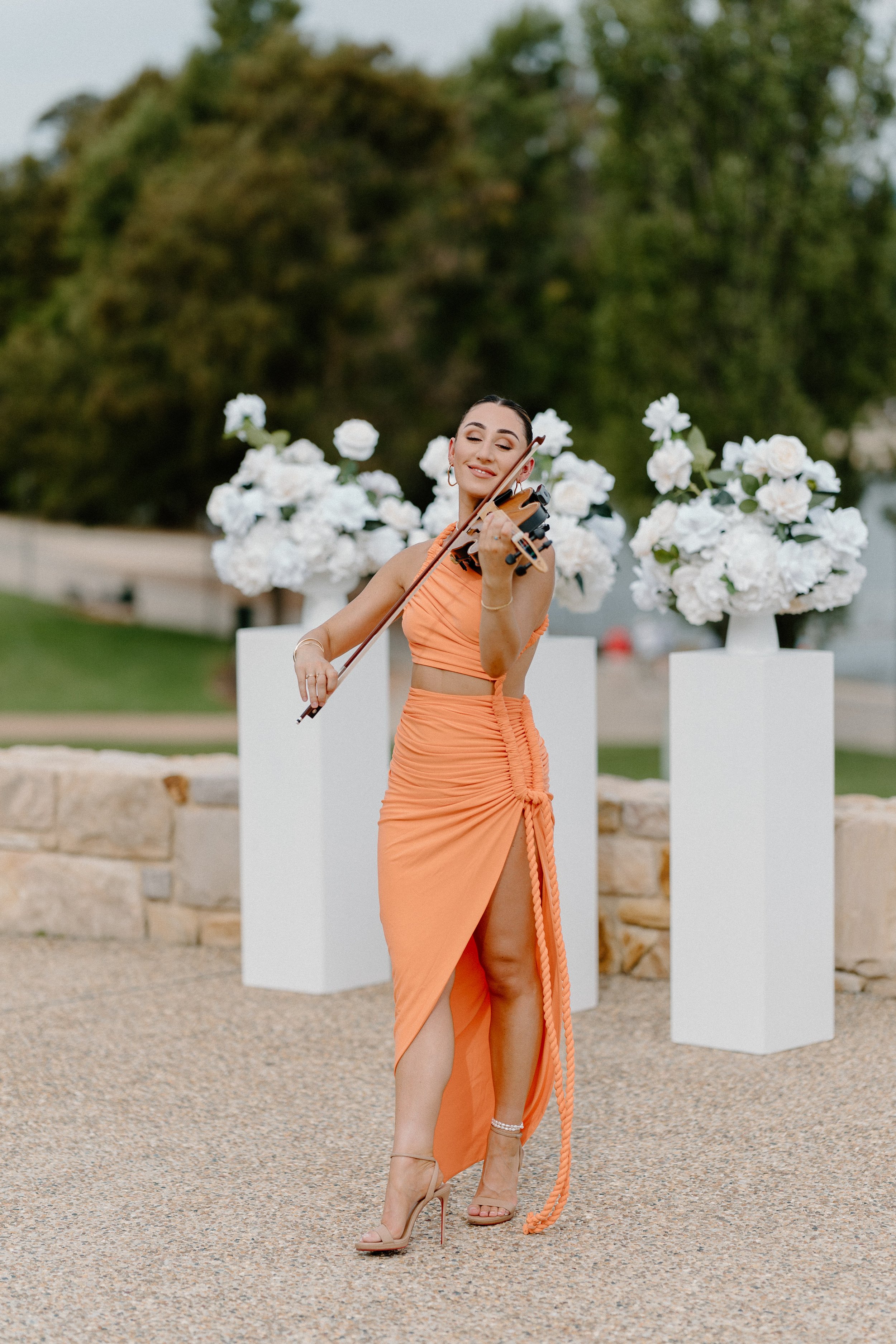 A woman in an elegant orange dress playing a violin outdoors, surrounded by white floral arrangements on white pedestals.