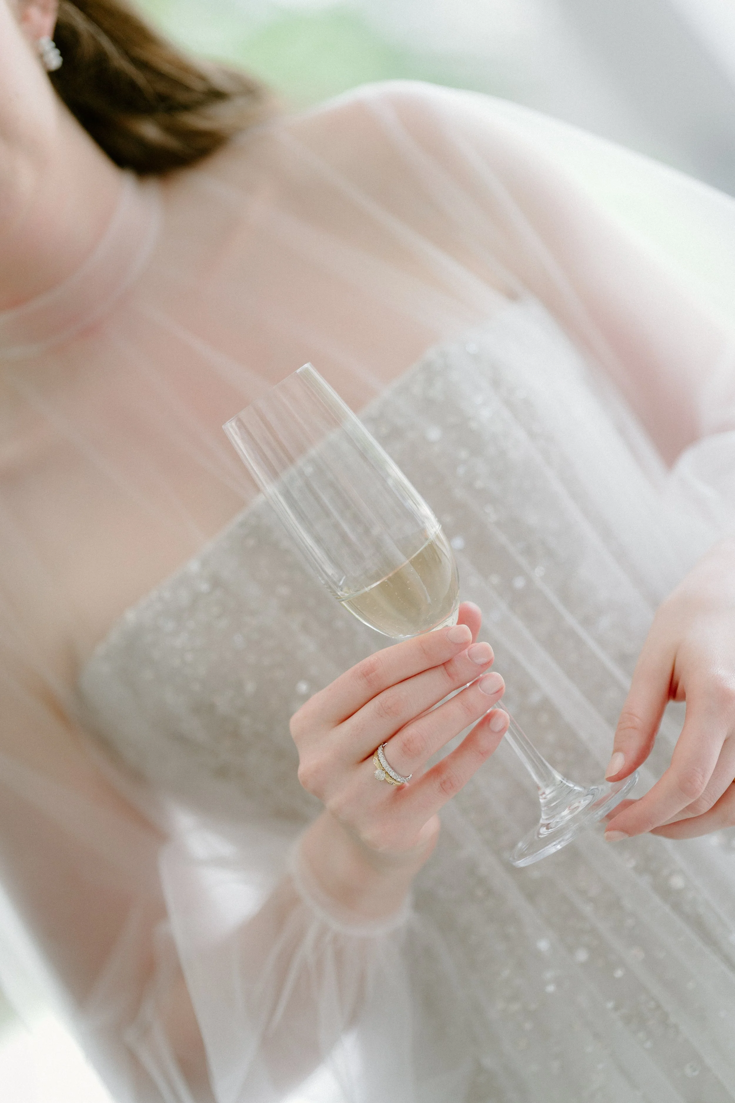Person holding a champagne flute with a light-colored beverage, wearing a wedding dress and a ring on their finger.