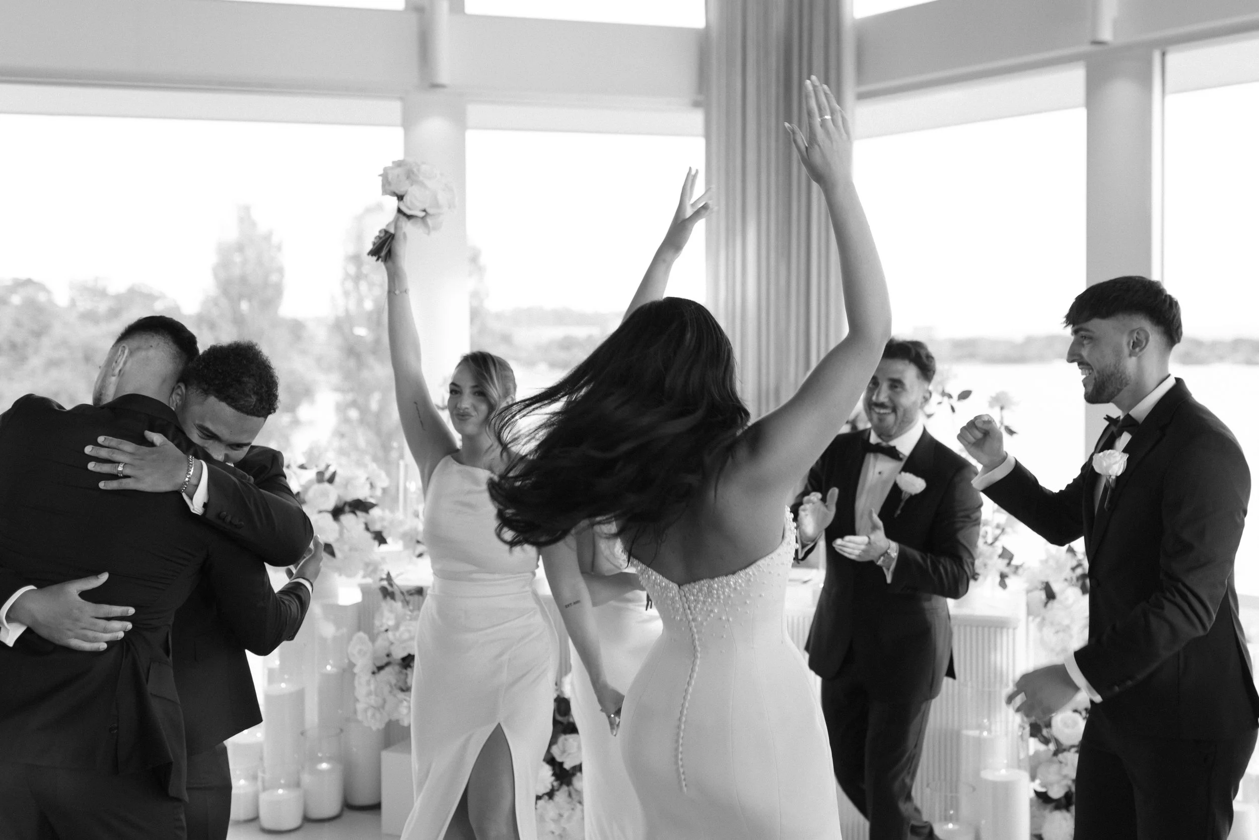 People celebrating at a wedding reception, hugging, dancing, and smiling indoors with large windows showing a scenic view outside.