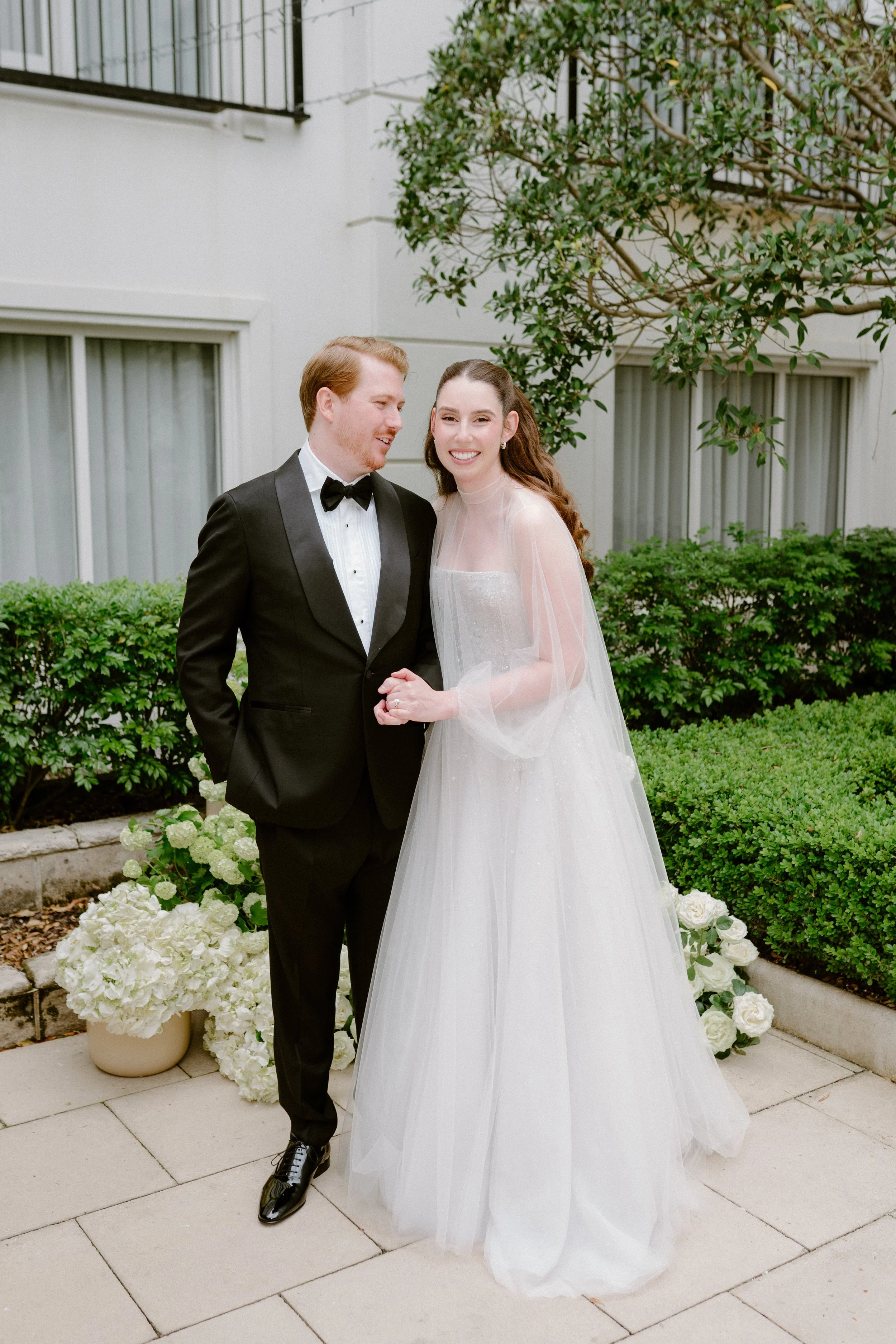 A bride and groom standing together outdoors on their wedding day, smiling. The groom is dressed in a black tuxedo with a bow tie, and the bride is wearing a white wedding gown with sheer sleeves and a tulle skirt, holding hands. There are plants and