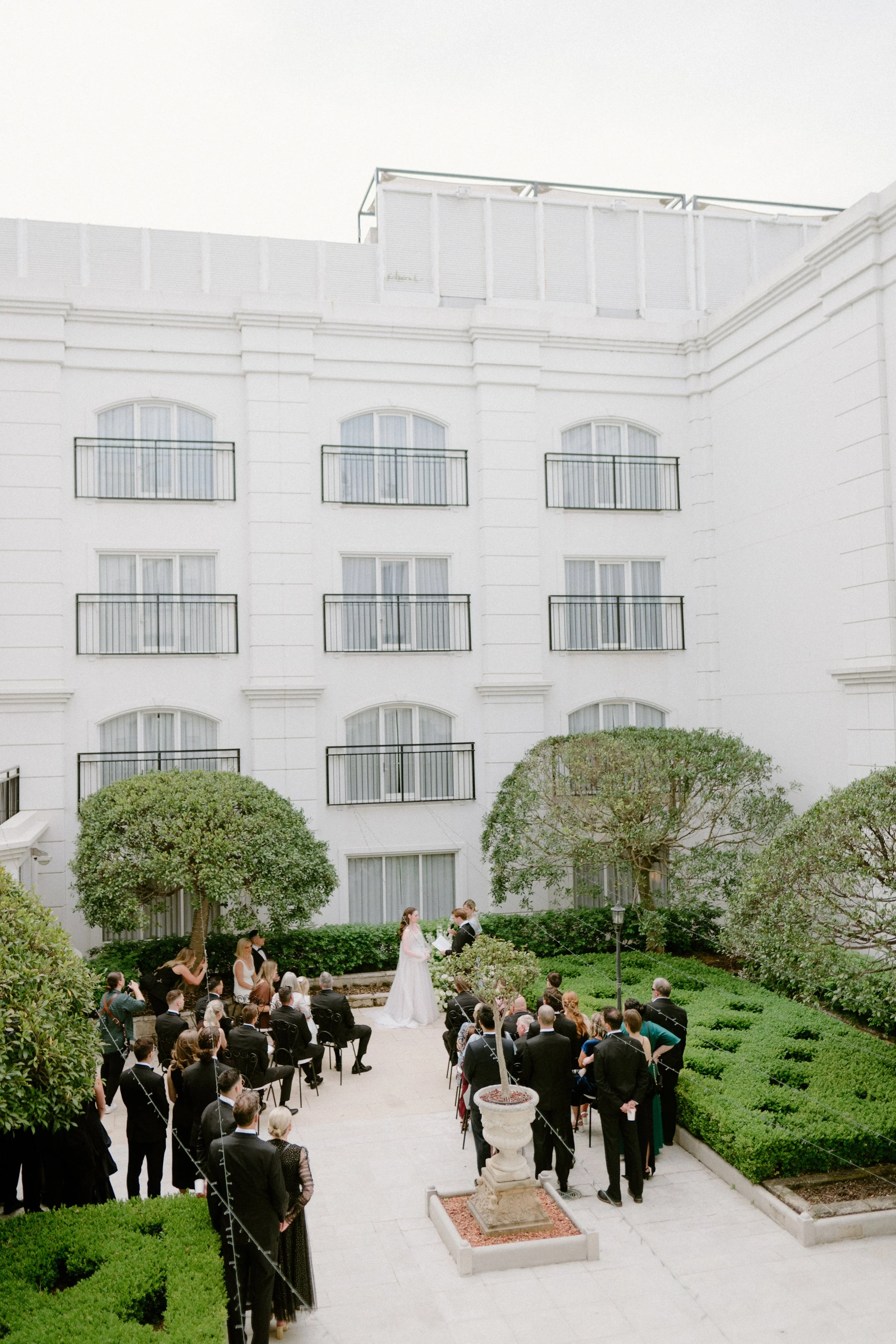 Outdoor wedding ceremony in a courtyard with a white building in the background, decorated with young trees and green bushes, with a bride and groom standing at the altar, surrounded by seated and standing guests.