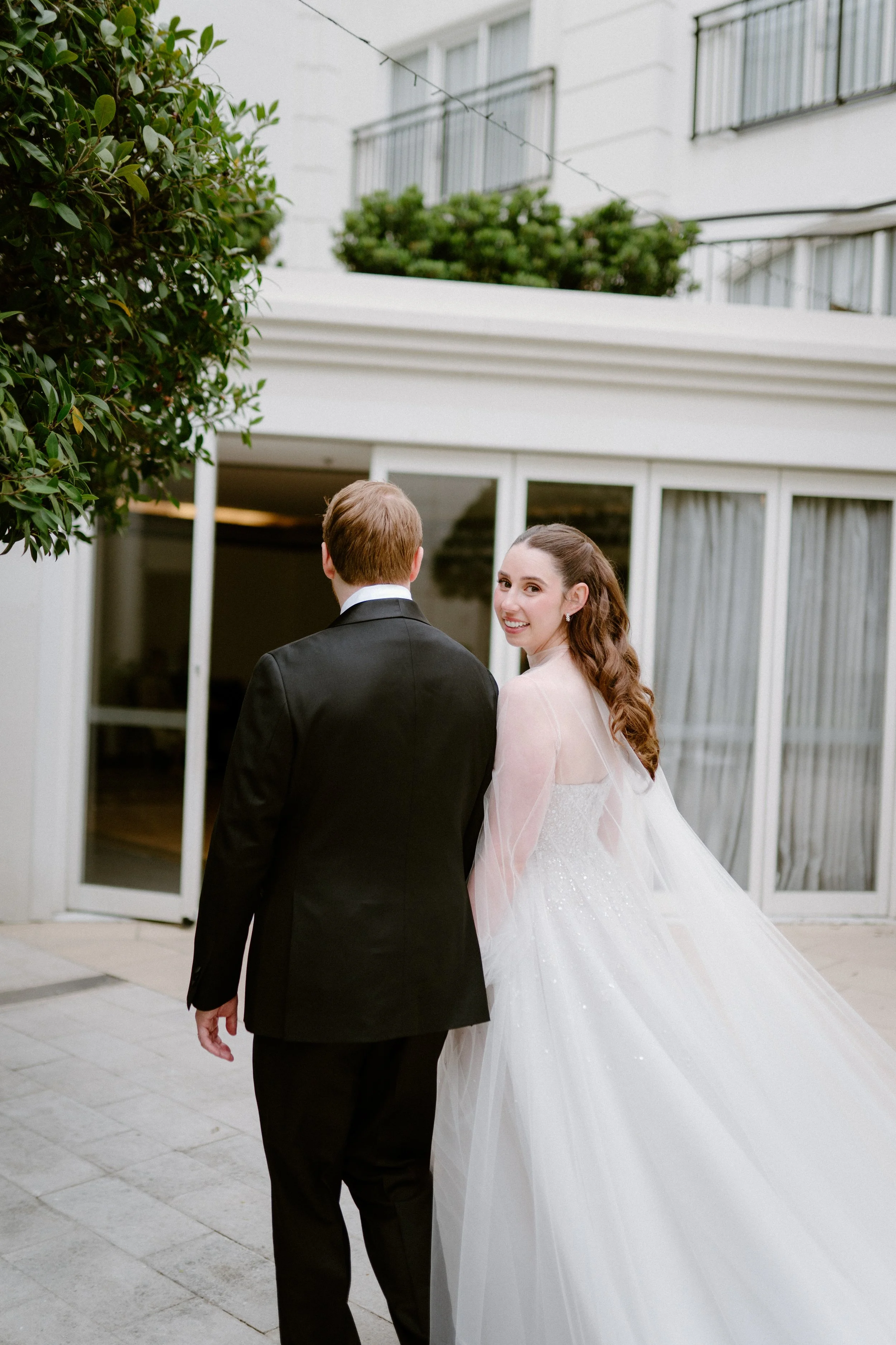 Bride in a white wedding dress and groom in a black suit walking outside near a building with glass doors, smiling back at the camera.
