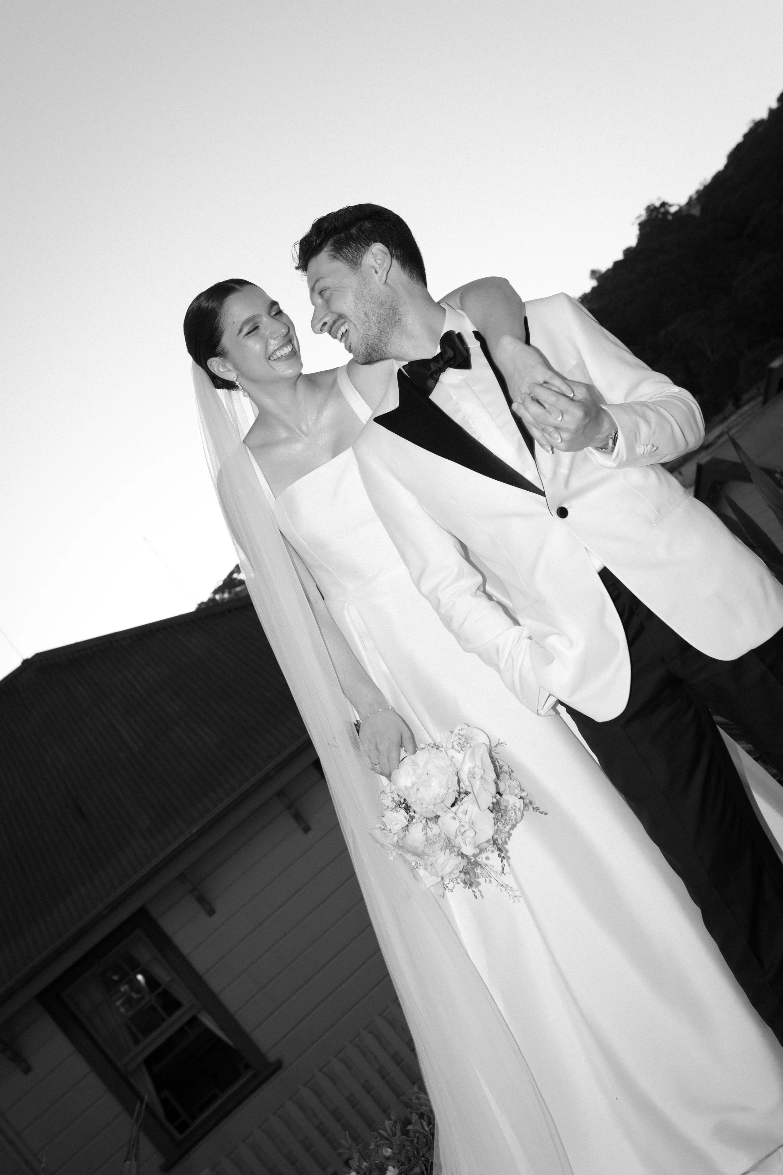 Sergeants Mess, Mosman Wedding Venue. Sergeants Mess Wedding Photos. A smiling bride in a wedding dress and veil holding a bouquet, standing next to a groom in a tuxedo, both laughing and leaning towards each other outdoors during wedding celebration