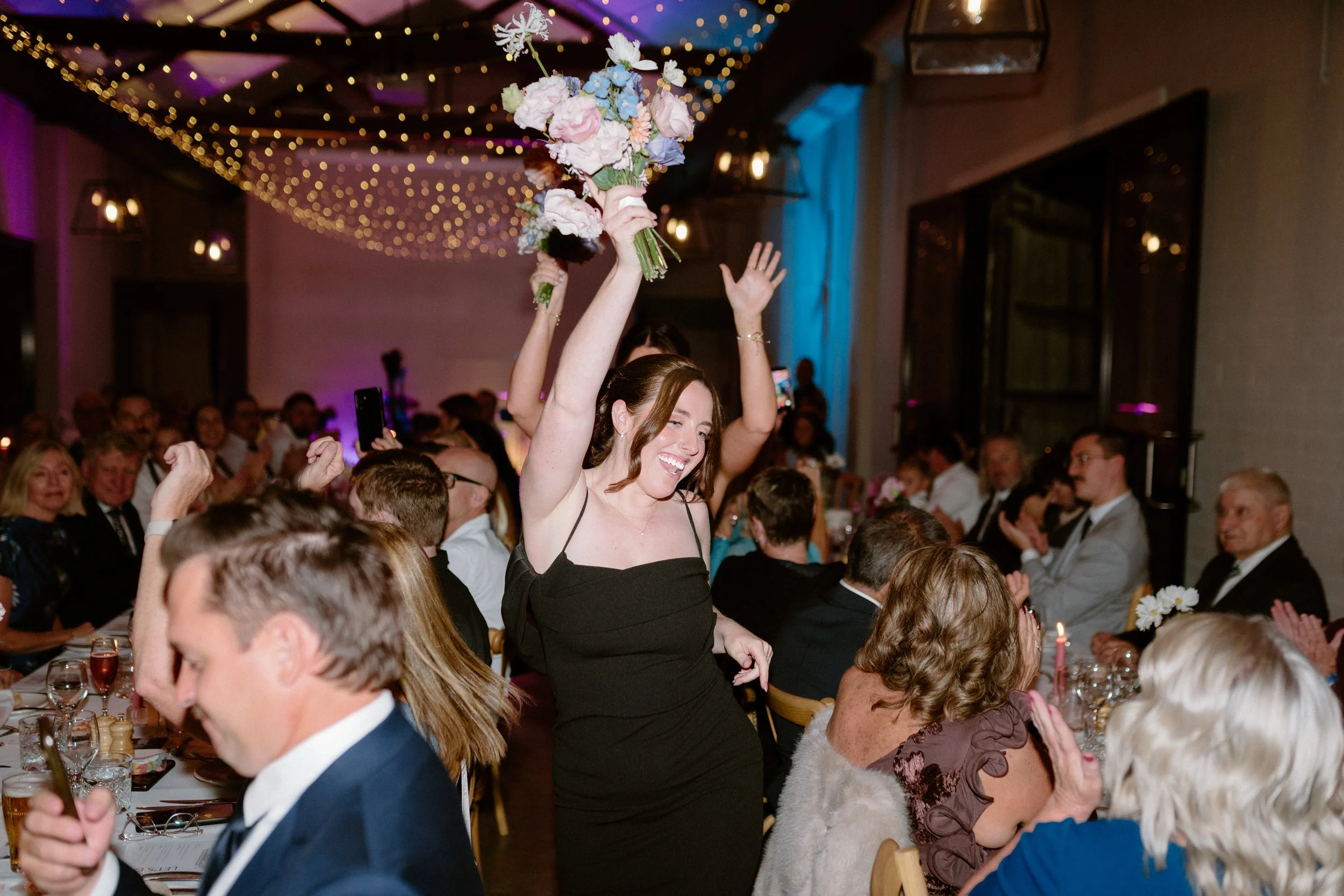 A woman in a black dress holding a large bouquet of pink and blue flowers at a crowded celebration or party. The room is decorated with string lights, and multiple guests are seated at tables, engaged in conversation or taking photos.