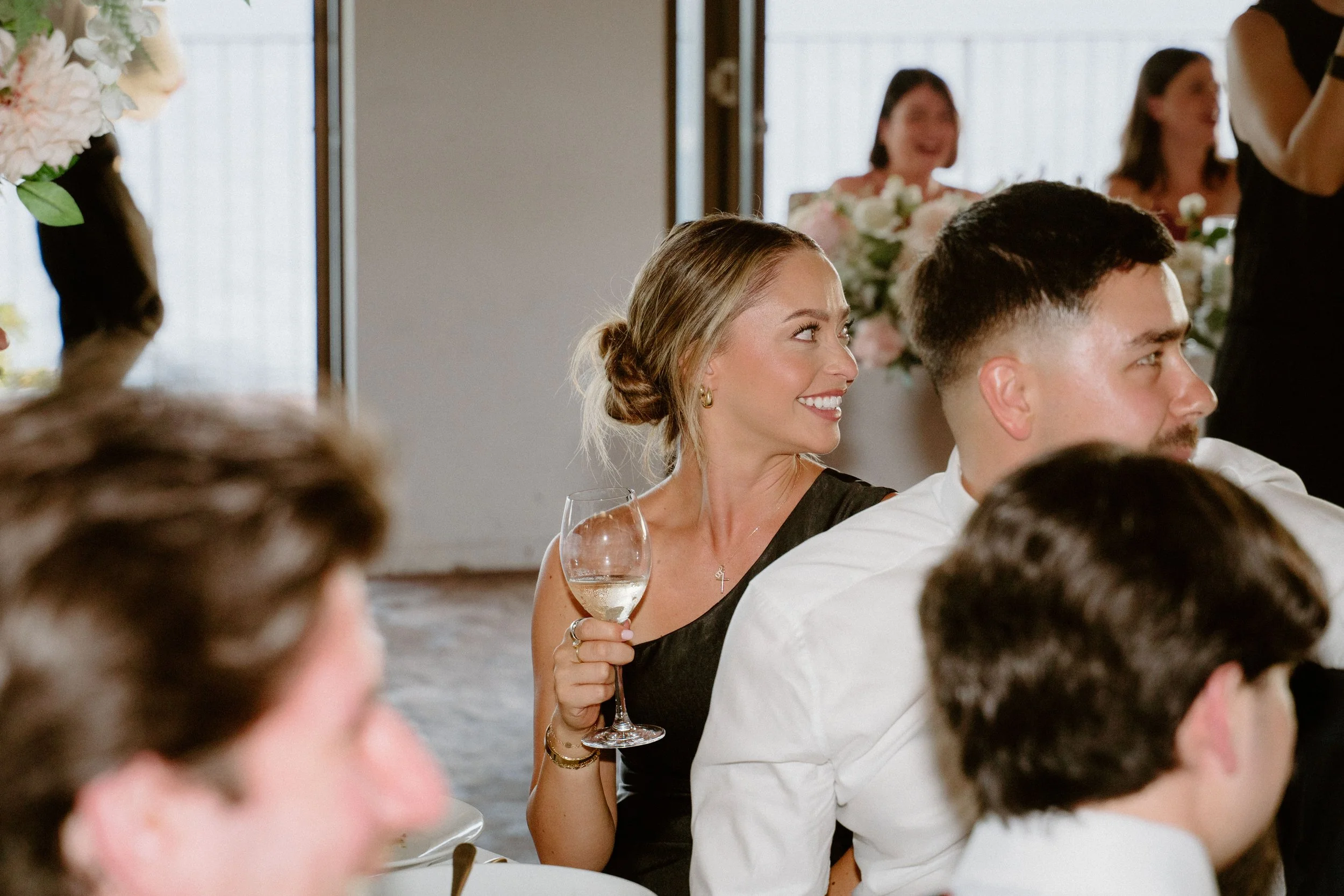 A woman with blond hair in an updo, wearing a black dress and earrings, holding a glass of white wine at a wedding reception, smiling and looking to the side. Other guests are visible around her, including a man with dark hair and a white shirt.
