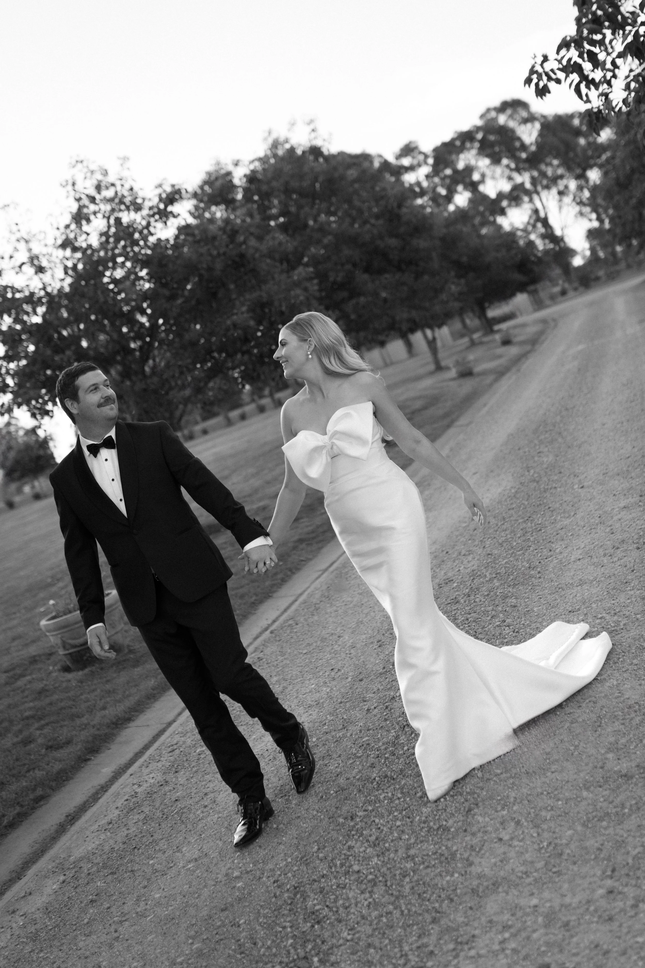 Black and white photo of a bride and groom holding hands and walking on a path in a park, with trees in the background.