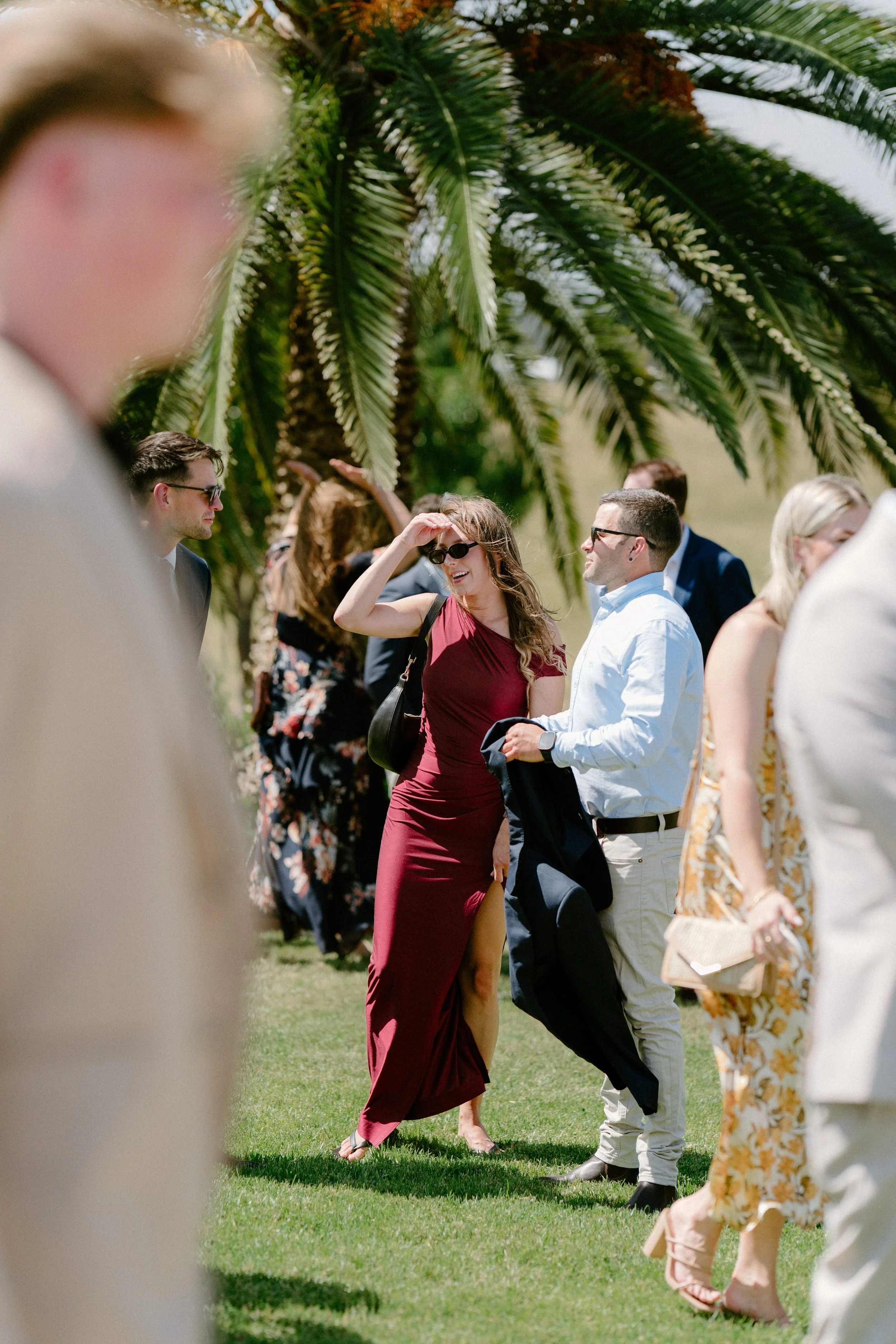 People dressed in semi-formal attire standing outdoors on grass, with a large palm tree in the background, enjoying sunny weather.