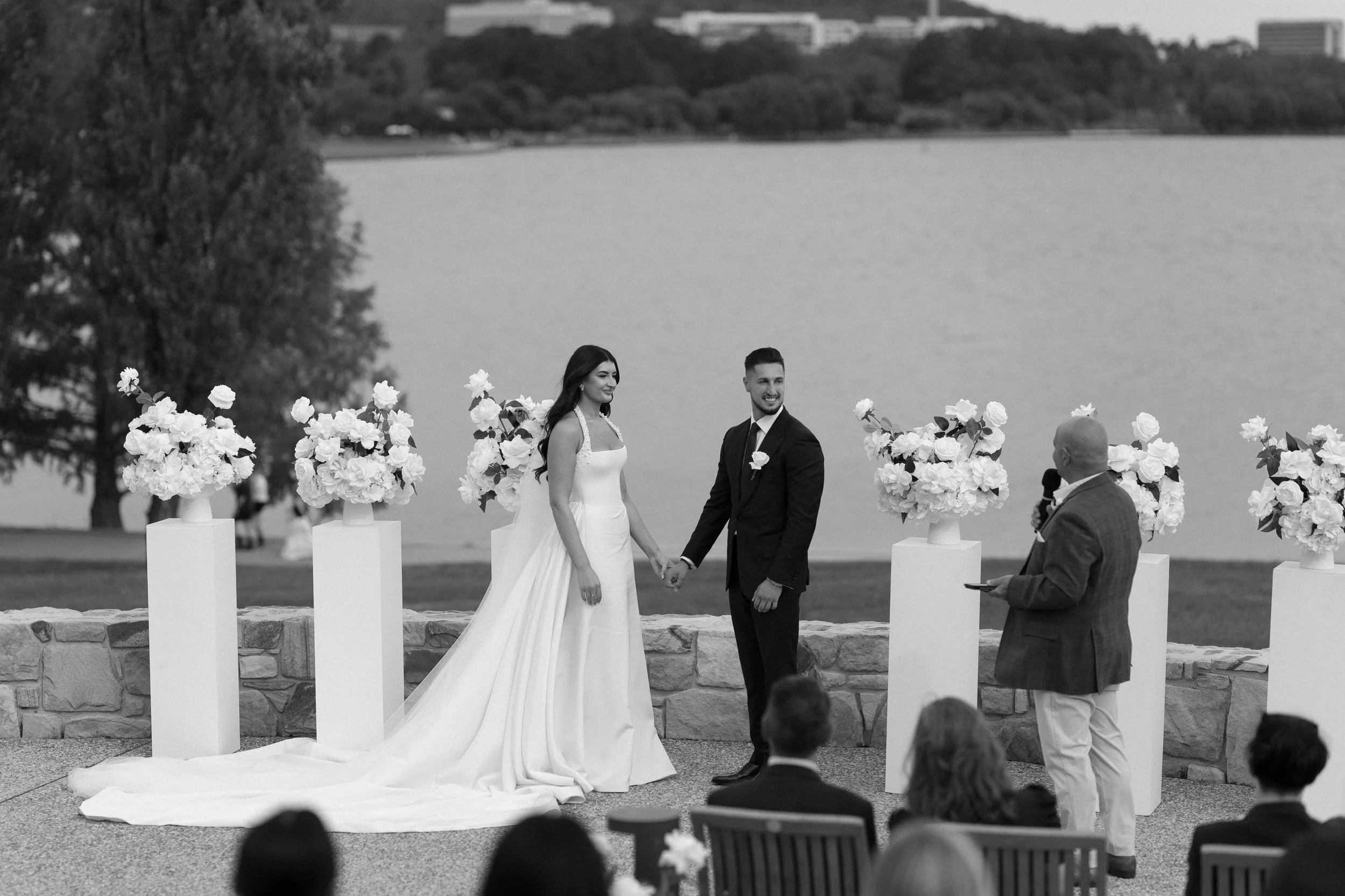 A black-and-white photo of a wedding ceremony by a lake. The bride and groom are holding hands and smiling, with the officiant speaking to them. The bride is wearing a long white wedding gown, and the groom is in a dark suit. There are six flower arr