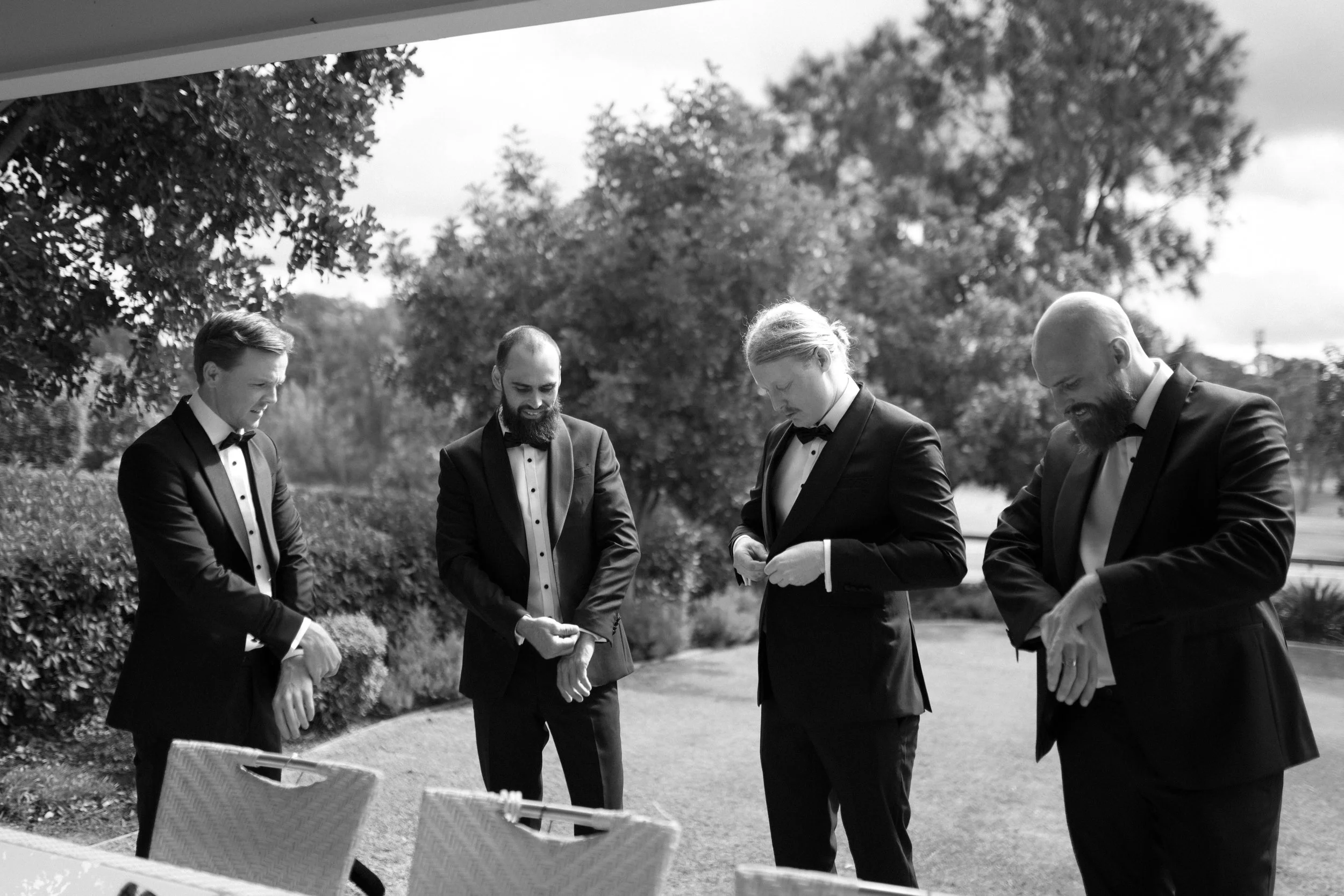 Four men in tuxedos standing outdoors with trees in the background, appearing to be engaged in a conversation or prayer.