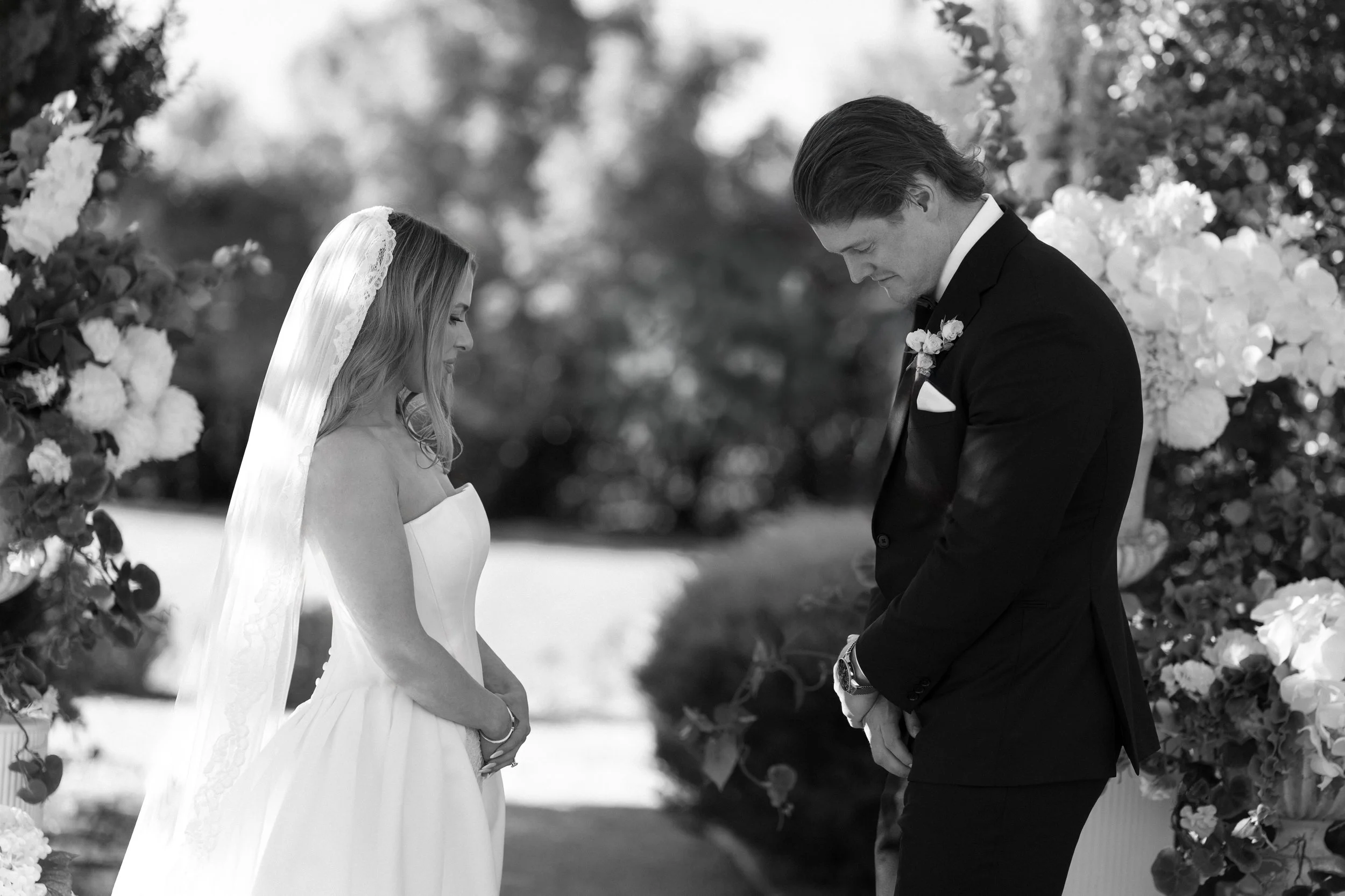 A black and white photograph of a bride and groom during a wedding ceremony outdoors, standing close with their heads bowed and hands clasped, surrounded by floral decorations.