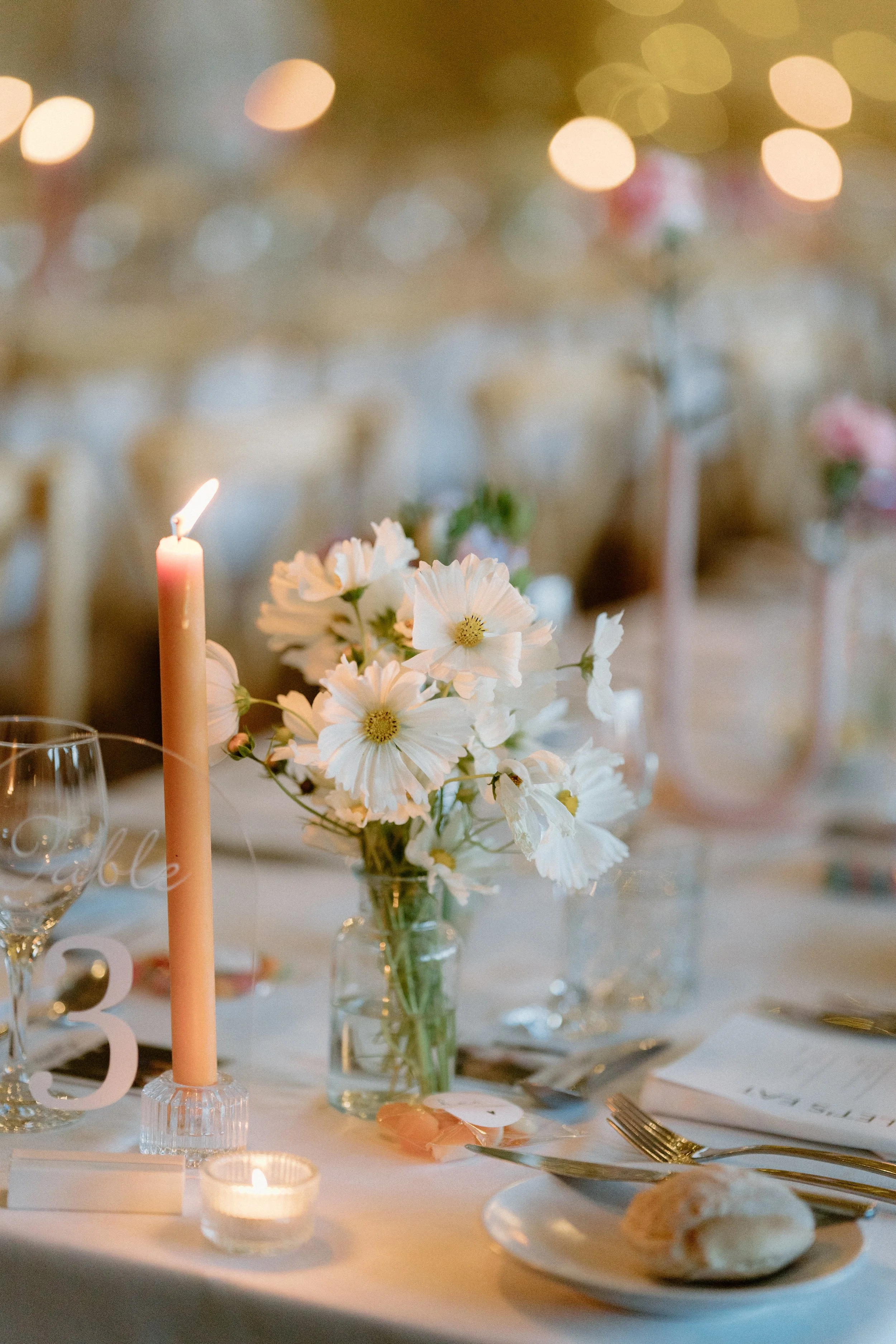 A decorated table with a pink candle, a floral centerpiece with white flowers, a lit candle, wine glasses, a plate with bread rolls, a white napkin, and gold-colored cutlery, set in a warmly lit venue with round bokeh lights in the background.