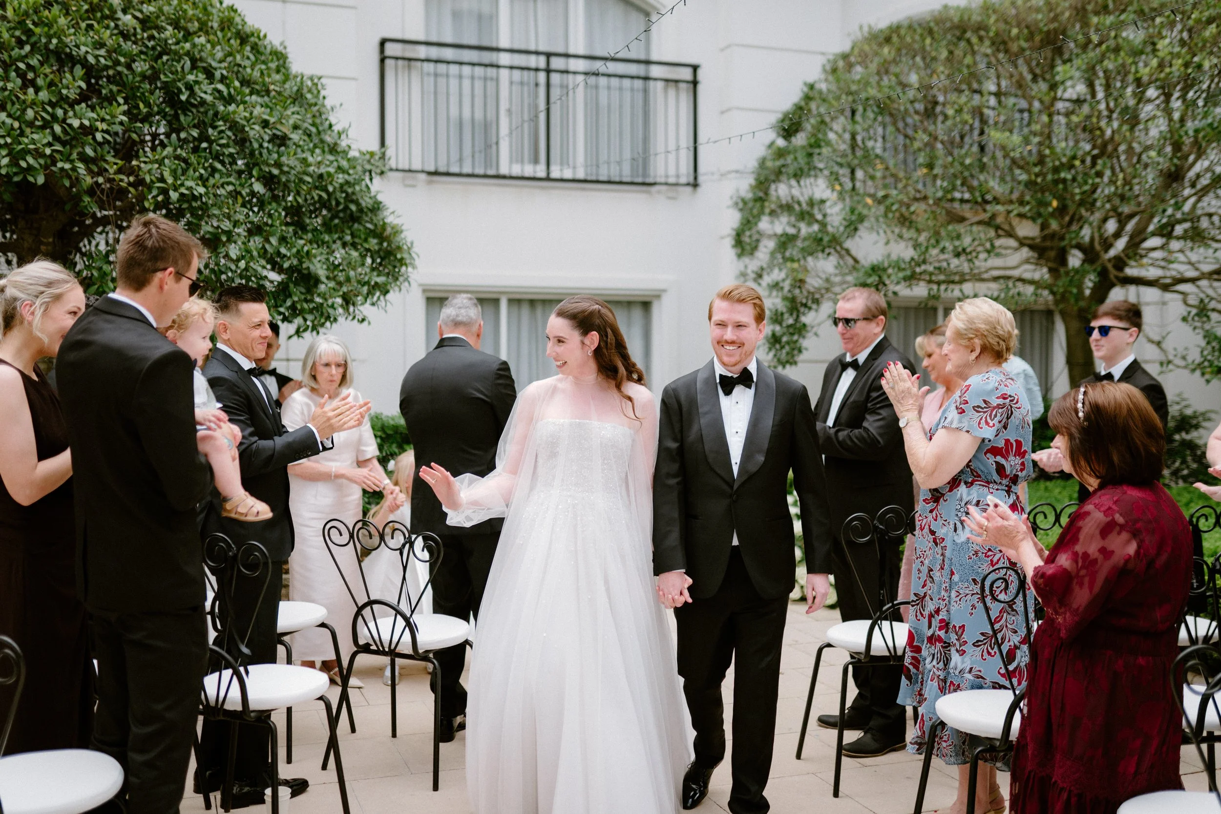A bride and groom walking hand in hand at their wedding reception outdoors, surrounded by friends and family clapping and smiling.