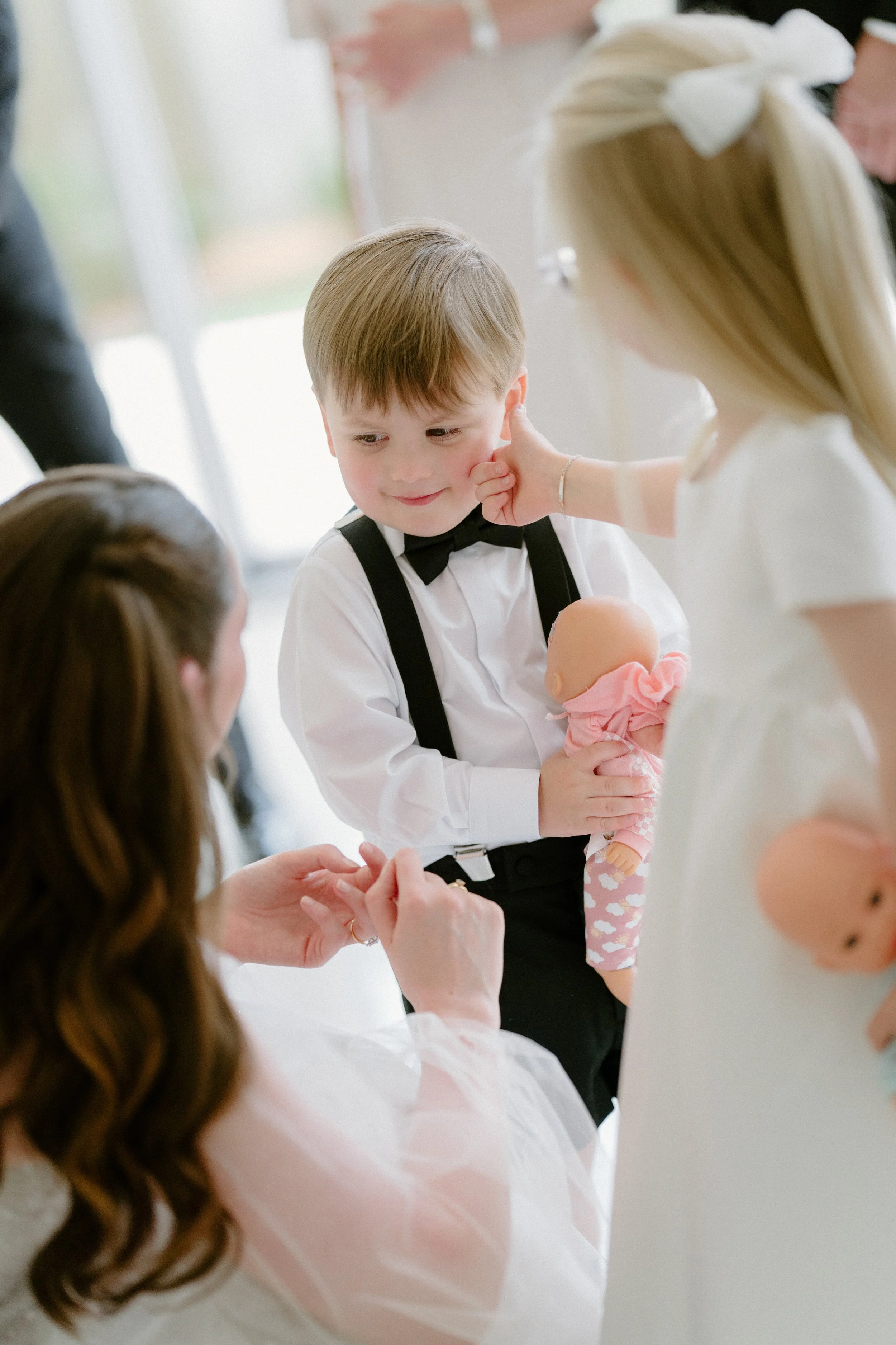 A young boy dressed in a white shirt with a black bow tie and suspenders is holding a doll and sitting at a gathering. A young girl with a large white bow in her hair is touching his face, and a woman with long brown hair is holding his hand, engagin
