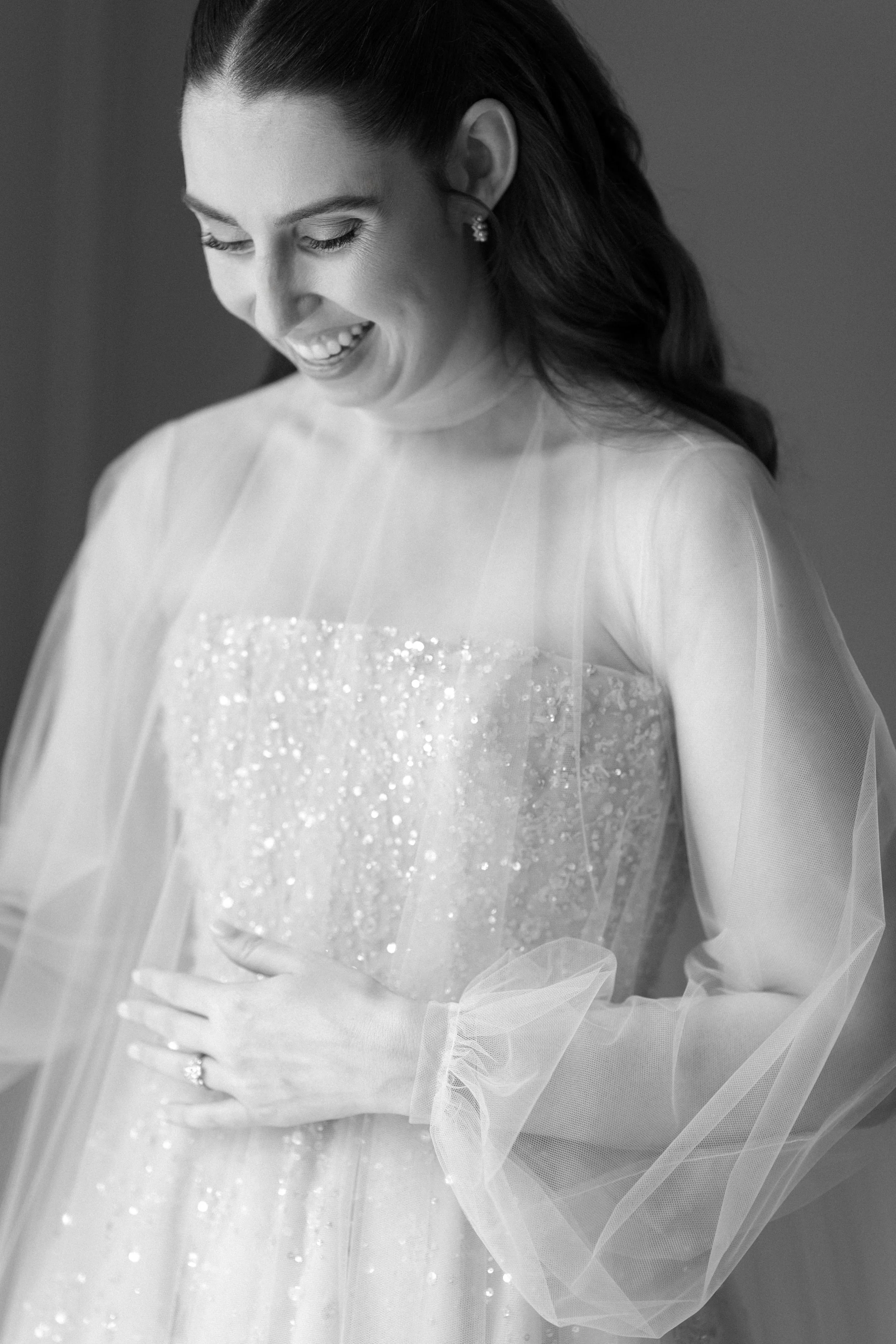 Black and white photo of a smiling woman in a wedding dress with a beaded bodice and sheer sleeves, looking down with her hand on her stomach.