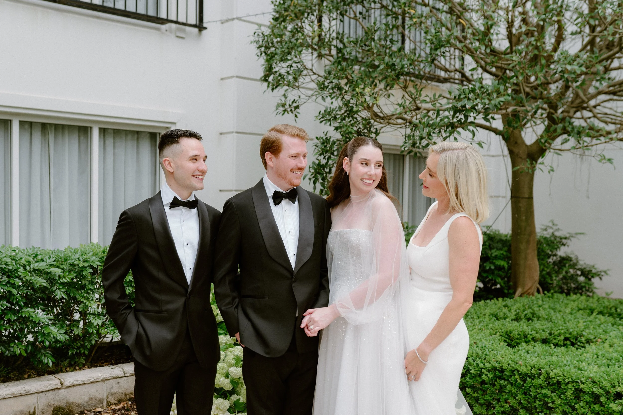 A wedding scene with three men in tuxedos and a woman in a white dress, standing outdoors near greenery, smiling and looking at a woman in a white gown.