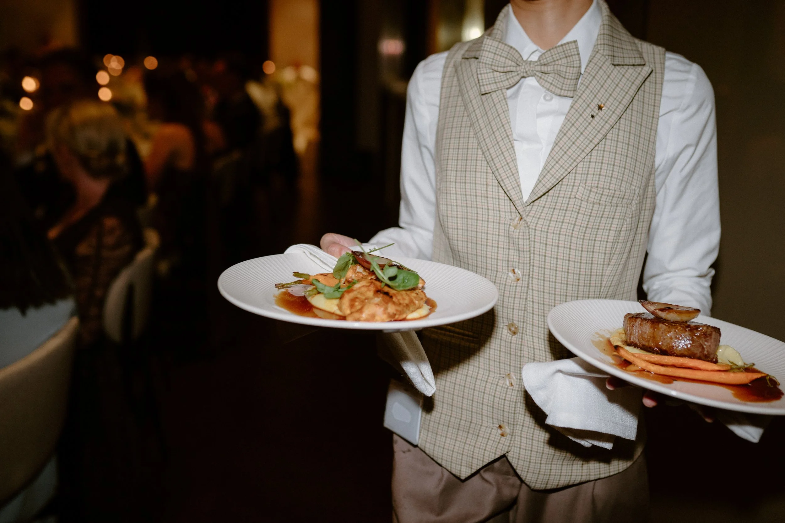 A waiter in a beige checkered vest and bow tie holds two plates of elegantly plated dishes at a formal dining event.