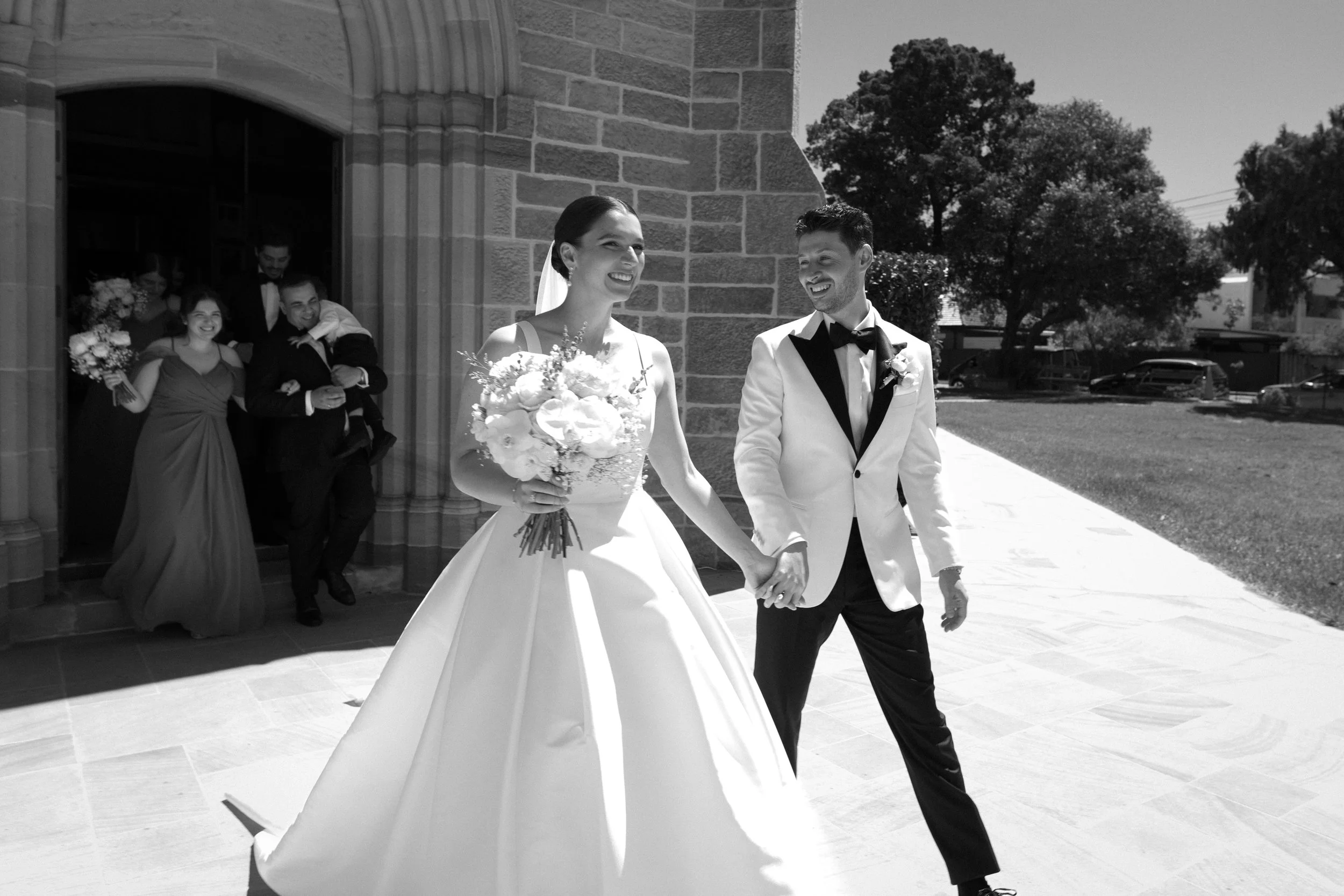 A wedding couple walking hand in hand outside a church, with bridesmaids and groomsmen in the background celebrating.