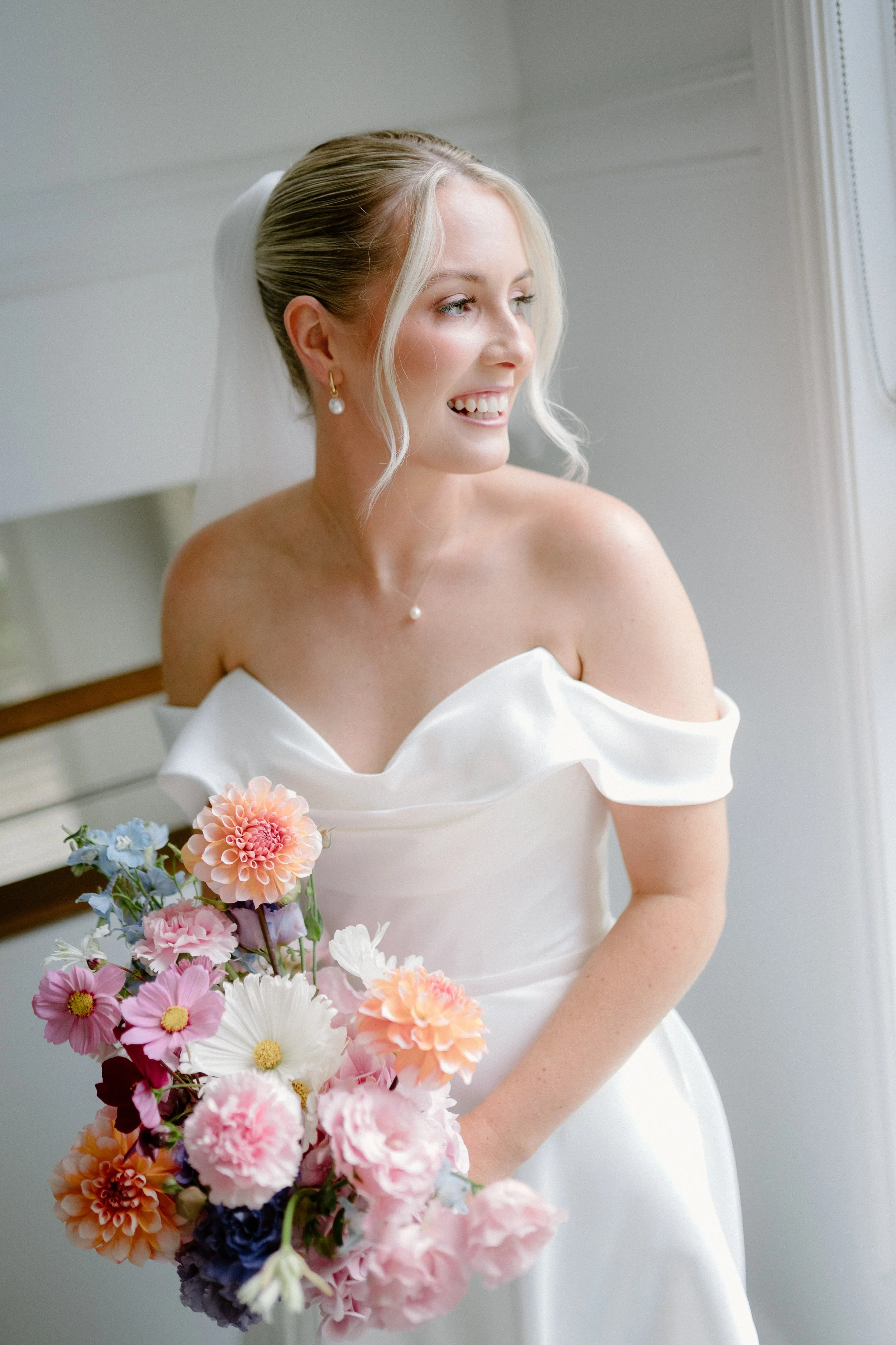 A woman in a white dress with off shoulder sleeves holding a colorful bouquet of flowers, smiling and looking to her right.