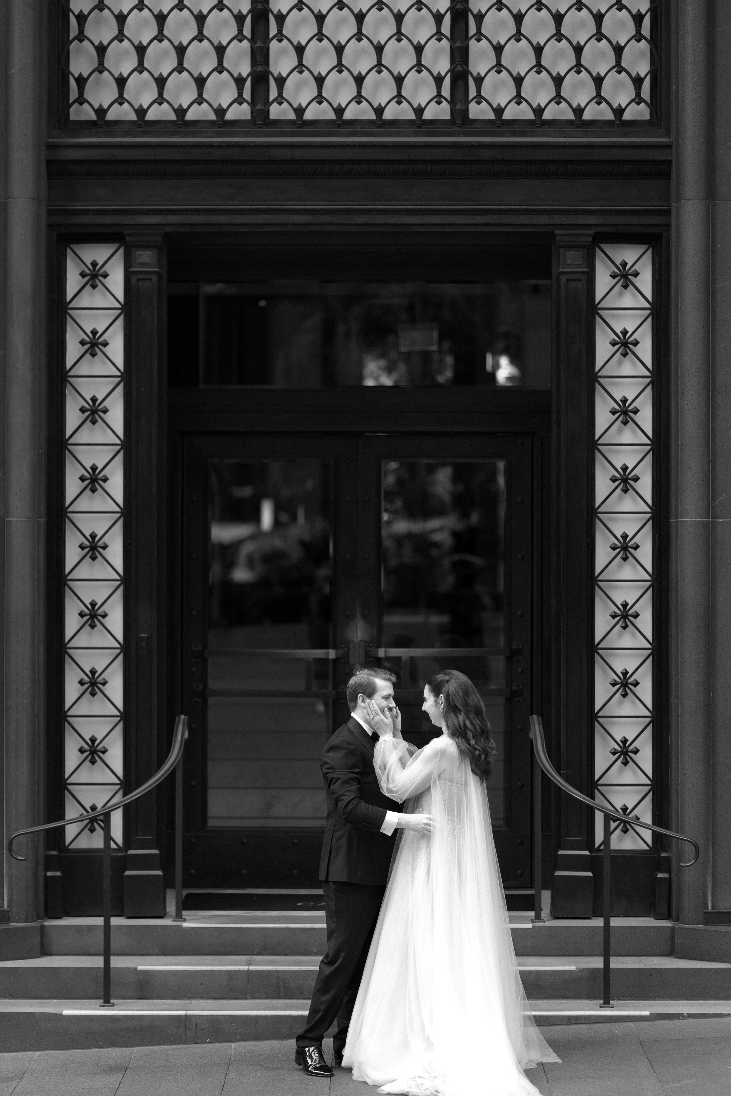 Black and white photo of a newlywed couple outside a building. The groom is in a black tuxedo, and the bride is in a long white wedding dress with sheer sleeves. They are holding hands and looking at each other, touching faces, standing on steps in f