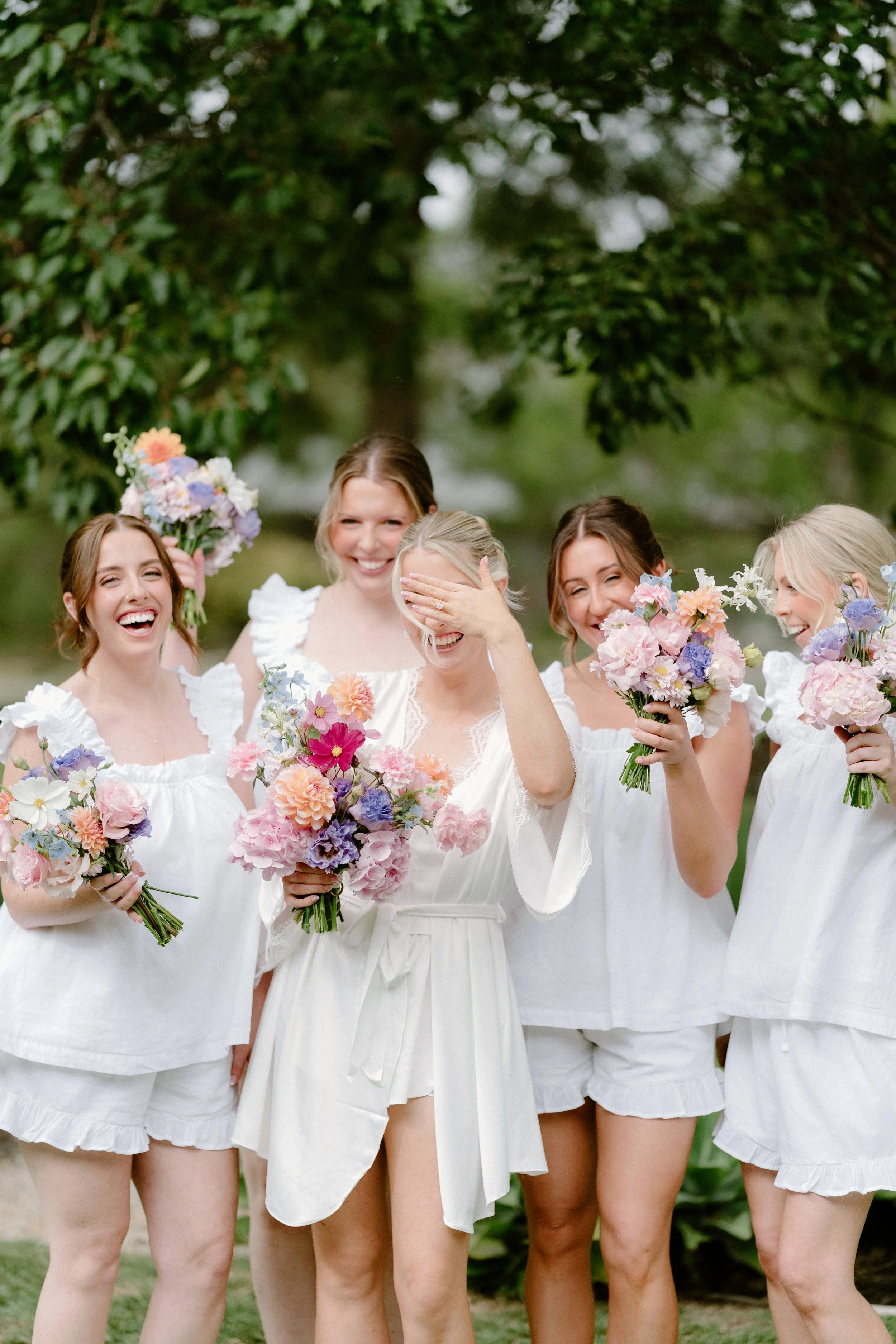 A group of women in white dresses holding colorful bouquets of flowers, standing outdoors under trees.
